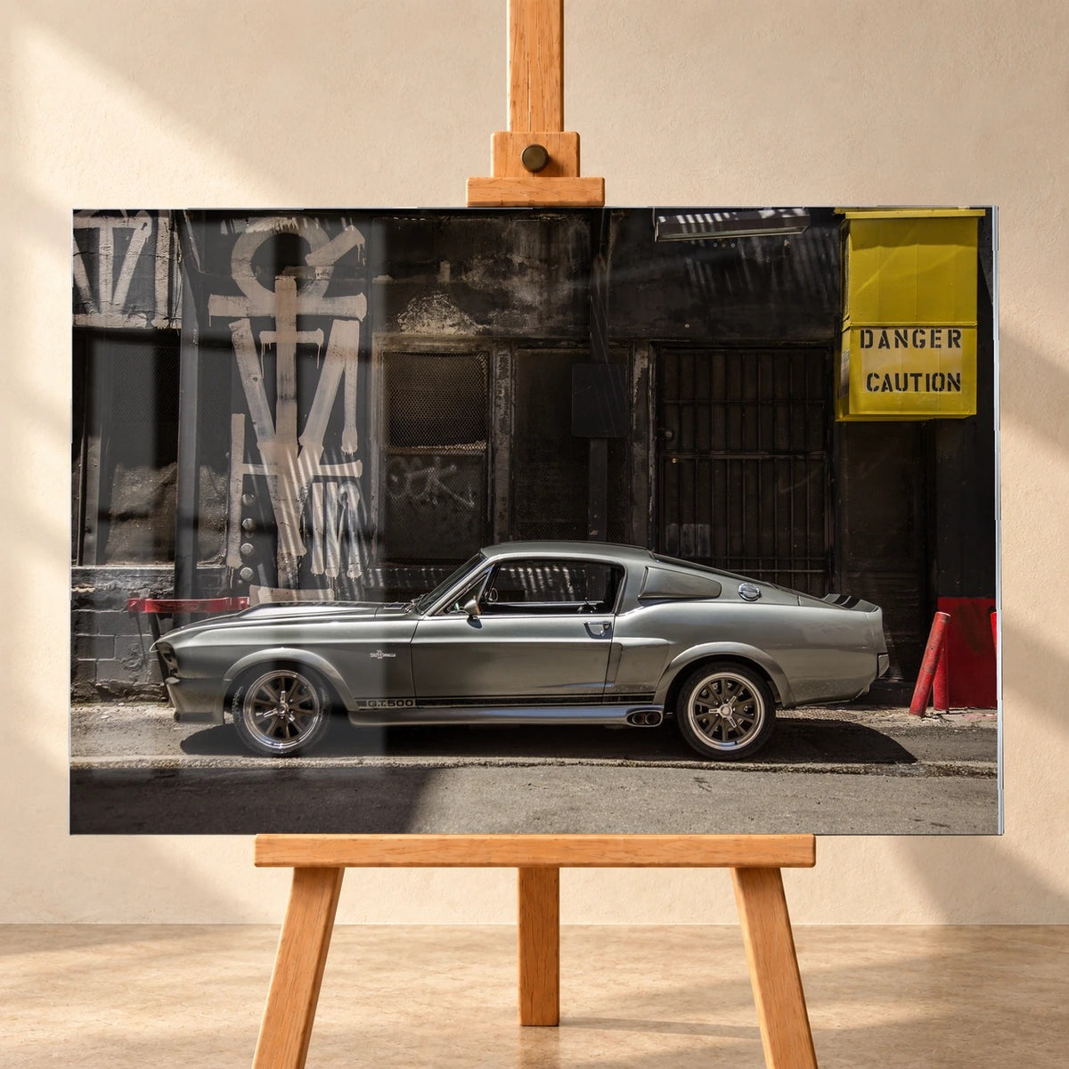 Frontal gezeigte Produktvisualisierung eines Acrylglas-Wandbilds auf einer Holzstaffelei vor heller Wand. Über dem Motiv des silbergrauen Ford Mustang Shelby GT500 „Eleanor“ vor einer dunklen Graffiti-Fassade sind Fensterreflexionen sichtbar.