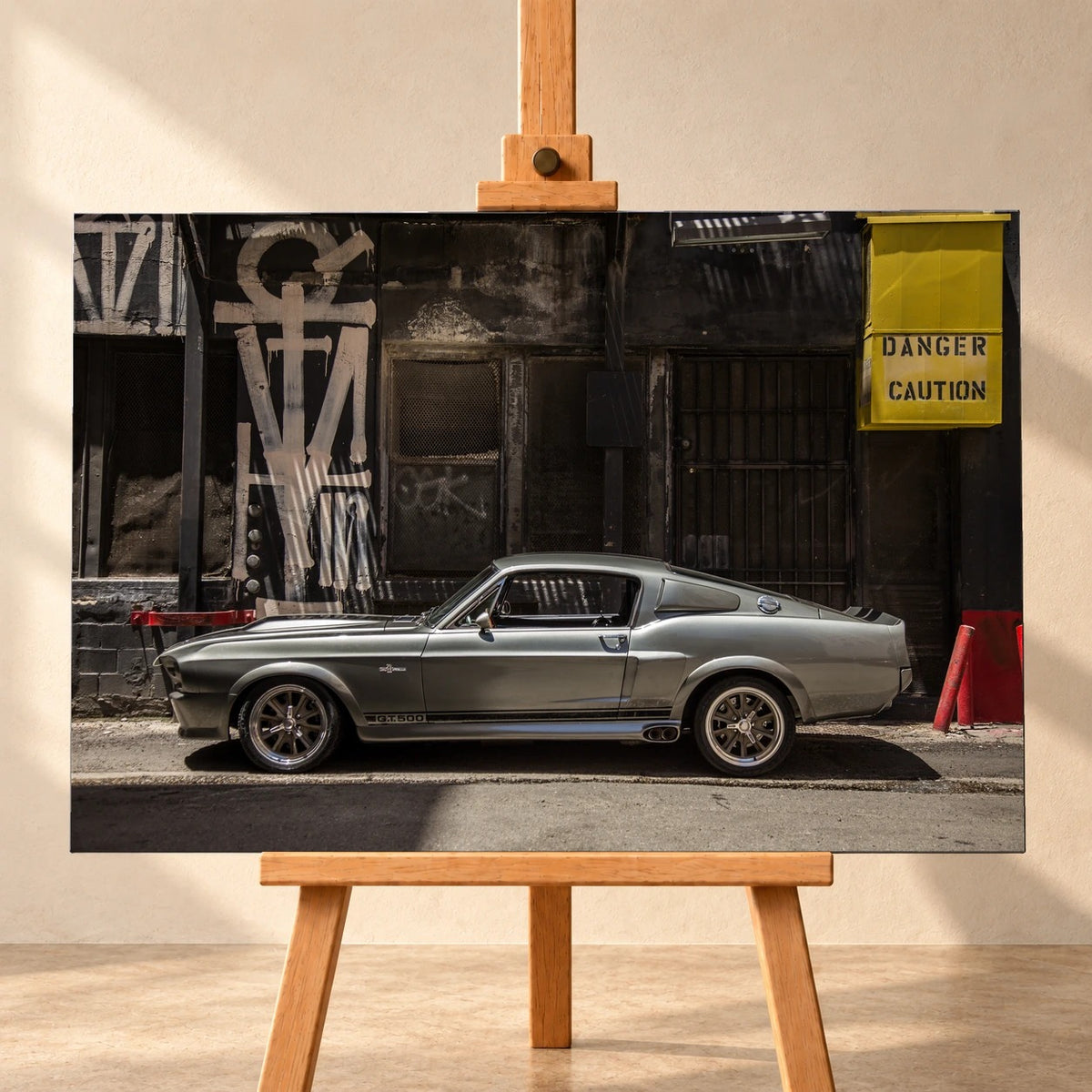 Visualisierung einer querformatigen Leinwand auf einer Holzstaffelei vor heller Wand. Das Motiv zeigt den silbergrauen Ford Mustang Shelby GT500 „Eleanor“ seitlich vor einer dunklen Industriefassade mit Graffiti und gelbem Warnkasten.