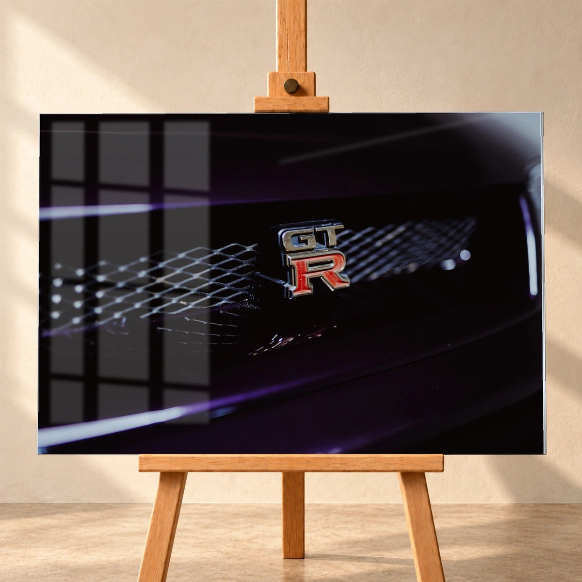 Frontal visualisiertes Acrylglas-Wandbild auf einer Holzstaffelei vor heller Wand. Über der linken Bildhälfte liegen Fensterreflexionen; das Motiv zeigt das silberne GT-R-Emblem mit rotem R am dunkelvioletten Kühlergrill des Nissan Skyline R33.