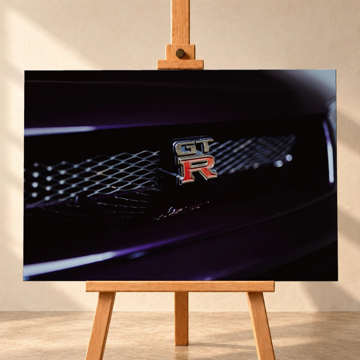 Visualisierung einer querformatigen Leinwand, frontal auf einer Holzstaffelei vor heller Wand. Das Motiv zeigt das silberne GT-R-Emblem mit rotem R am dunklen Kühlergrill eines Nissan Skyline R33, umgeben von violetten Lichtakzenten.