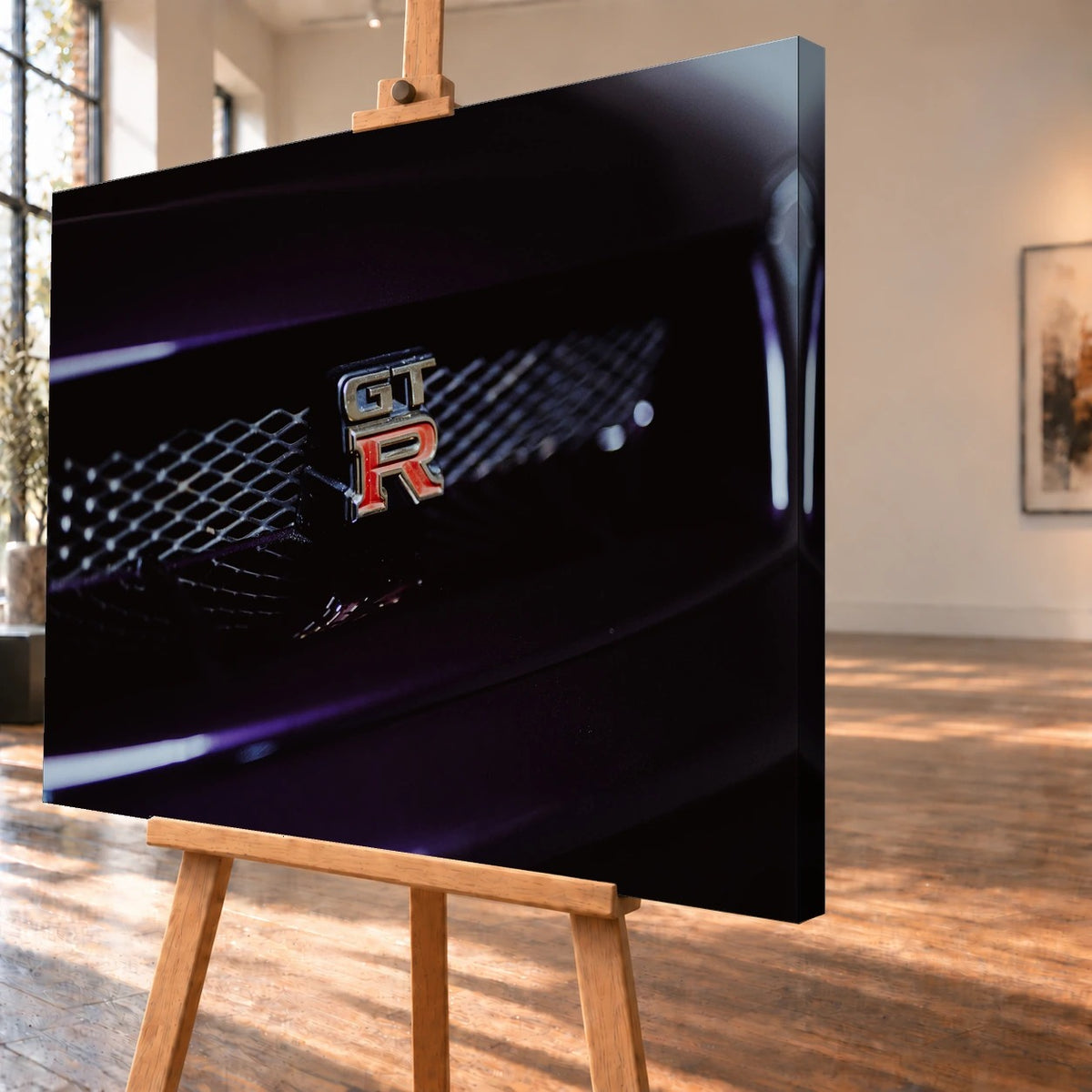 Schrägansicht einer Leinwand als Produktvisualisierung auf einer Holzstaffelei; die tiefe rechte Seitenkante ist sichtbar. Auf dem Motiv hebt sich das silberne GT-R-Emblem mit rotem R vom dunklen Gittergrill des Nissan Skyline R33 ab.