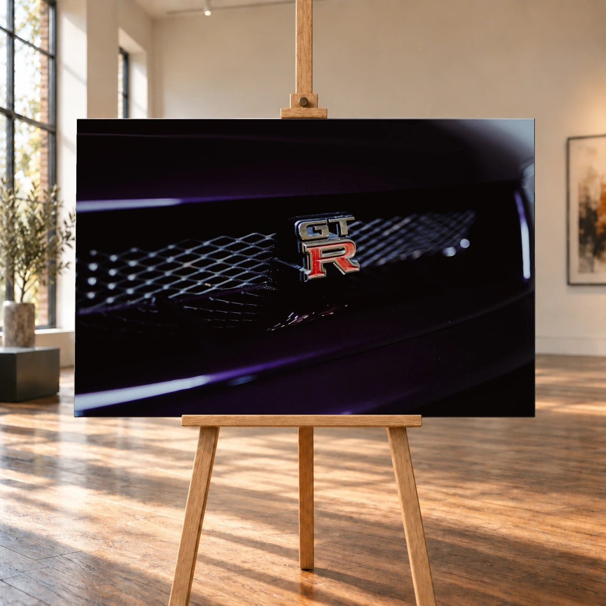 Raumvisualisierung mit querformatiger Leinwand auf einer Holzstaffelei in einem hellen Raum mit großen Fenstern und Holzboden. Die Leinwand zeigt eine Nahaufnahme des GT-R-Emblems am dunklen Kühlergrill eines Nissan Skyline R33.