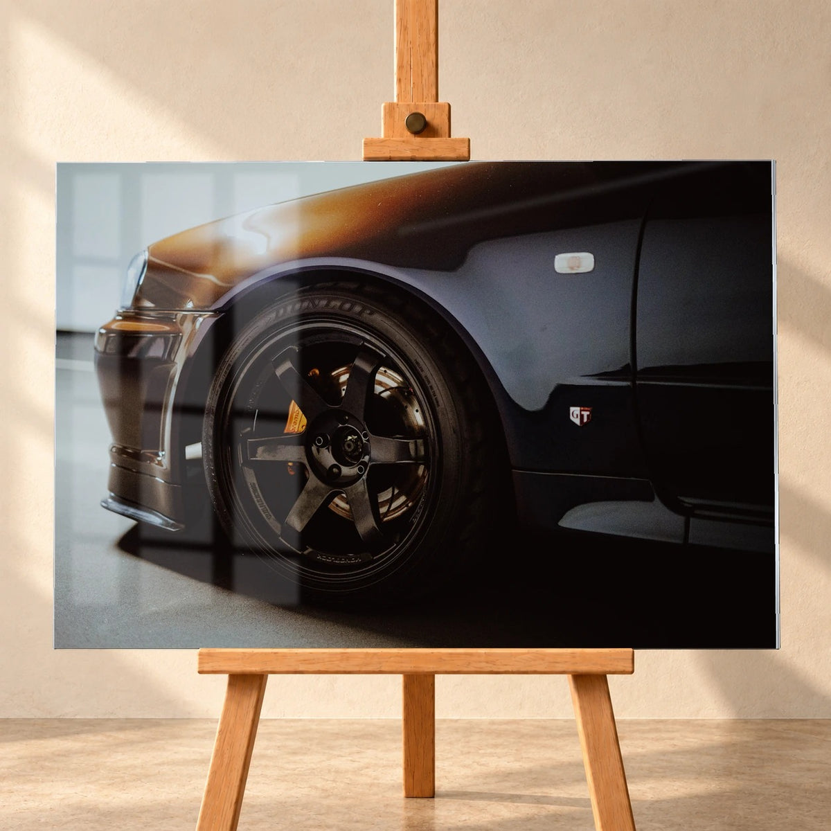 Visualisierung eines Acrylglas-Wandbilds im Querformat, frontal auf einer Holzstaffelei vor heller Wand. Dargestellte Fensterreflexionen überlagern das Motiv eines dunklen Nissan Skyline R34 mit schwarzem Vorderrad und gelbem Bremssattel.