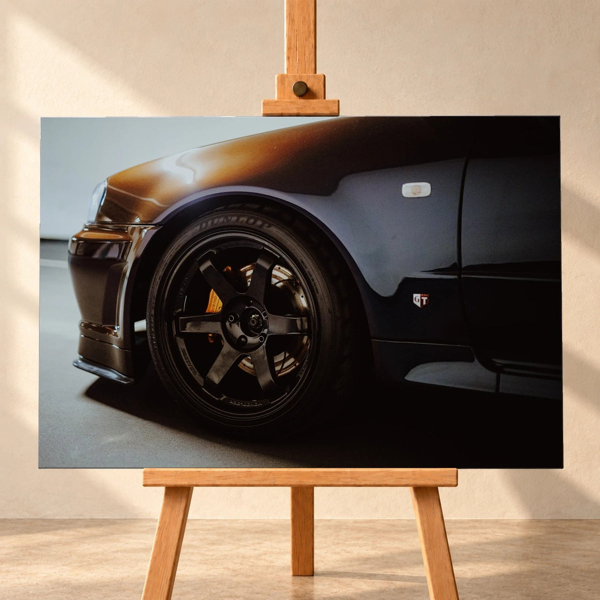 Frontale Visualisierung eines querformatigen Leinwandbilds auf einer Holzstaffelei vor heller Wand. Das Motiv zeigt Vorderrad und Kotflügel eines dunklen Nissan Skyline R34 mit schwarzer Sechsspeichenfelge und gelbem Bremssattel.