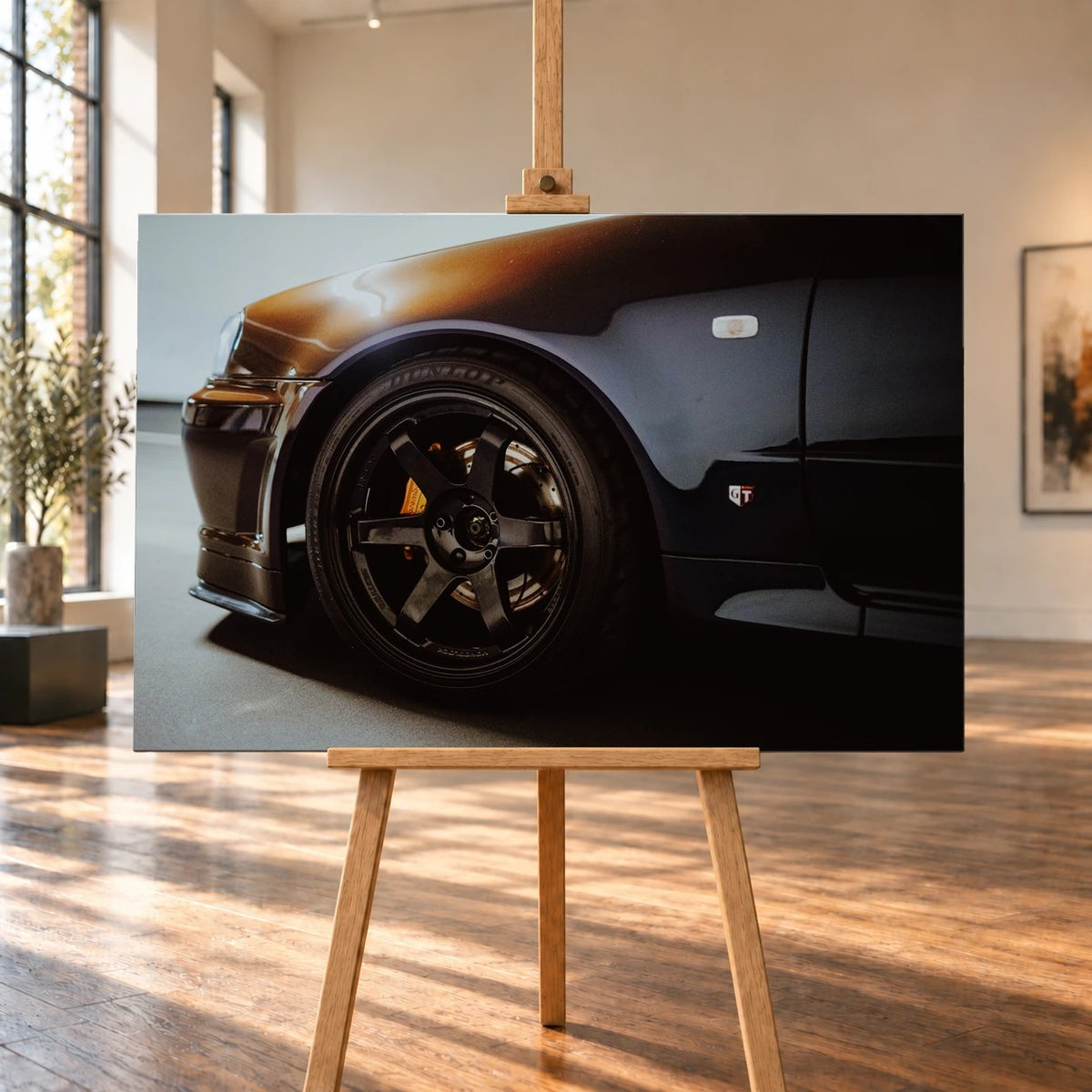 Visualisierte Raumansicht eines querformatigen Leinwandbilds auf einer Holzstaffelei in einem hellen Raum mit hohen Fenstern und Holzboden. Das Motiv zeigt das Vorderrad eines Nissan Skyline R34 und warme Lichtreflexe auf dem dunklen Kotflügel.