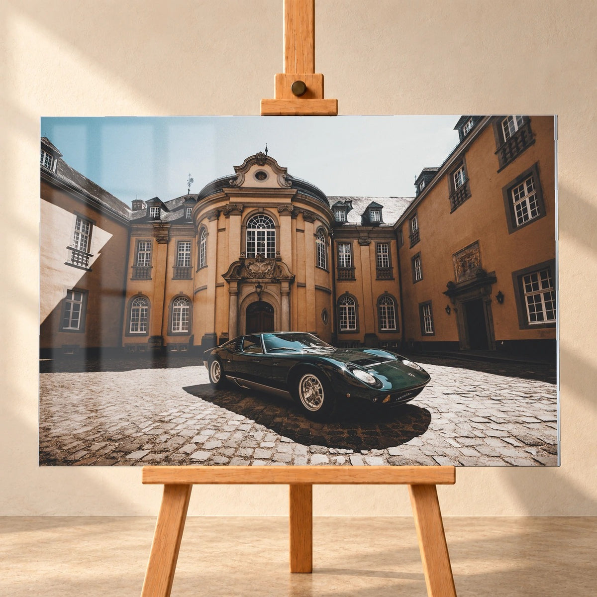 Frontal visualisiertes Acrylglasbild im Querformat auf einer Holzstaffelei vor heller Wand, mit sichtbaren Fensterspiegelungen. Im Motiv steht ein dunkelgrüner klassischer Sportwagen auf Kopfsteinpflaster vor einer ockerfarbenen Barockfassade.
