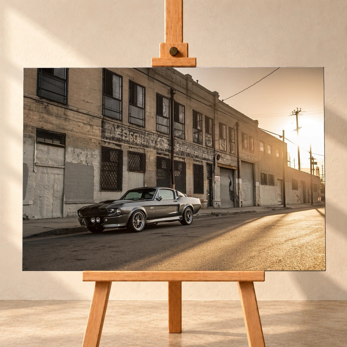 Visualisierung eines Leinwandbildes im Querformat auf einer Holzstaffelei vor heller Wand. Das Motiv zeigt den silbergrauen Ford Mustang Shelby GT500 „Eleanor“ vor einer verwitterten Industriefassade im tief stehenden Sonnenlicht.