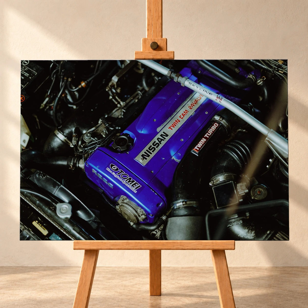 Visualisierung eines querformatigen Leinwandbilds auf einer Holzstaffelei vor heller Wand. Das Motiv zeigt den Motorraum eines Nissan Skyline R33 mit violettem Ventildeckel und diagonal verlaufender silberner Domstrebe.