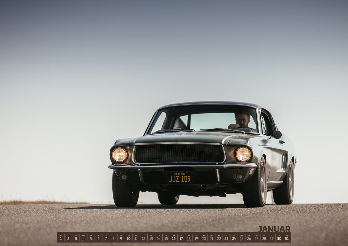 Bullitt Kalender 2027 – Januar: Dunkelgrüner klassischer Ford Mustang in tiefer Frontansicht auf Asphalt, mit eingeschalteten Rundscheinwerfern, sichtbarem Fahrer und weitem hellgrauem Himmel
