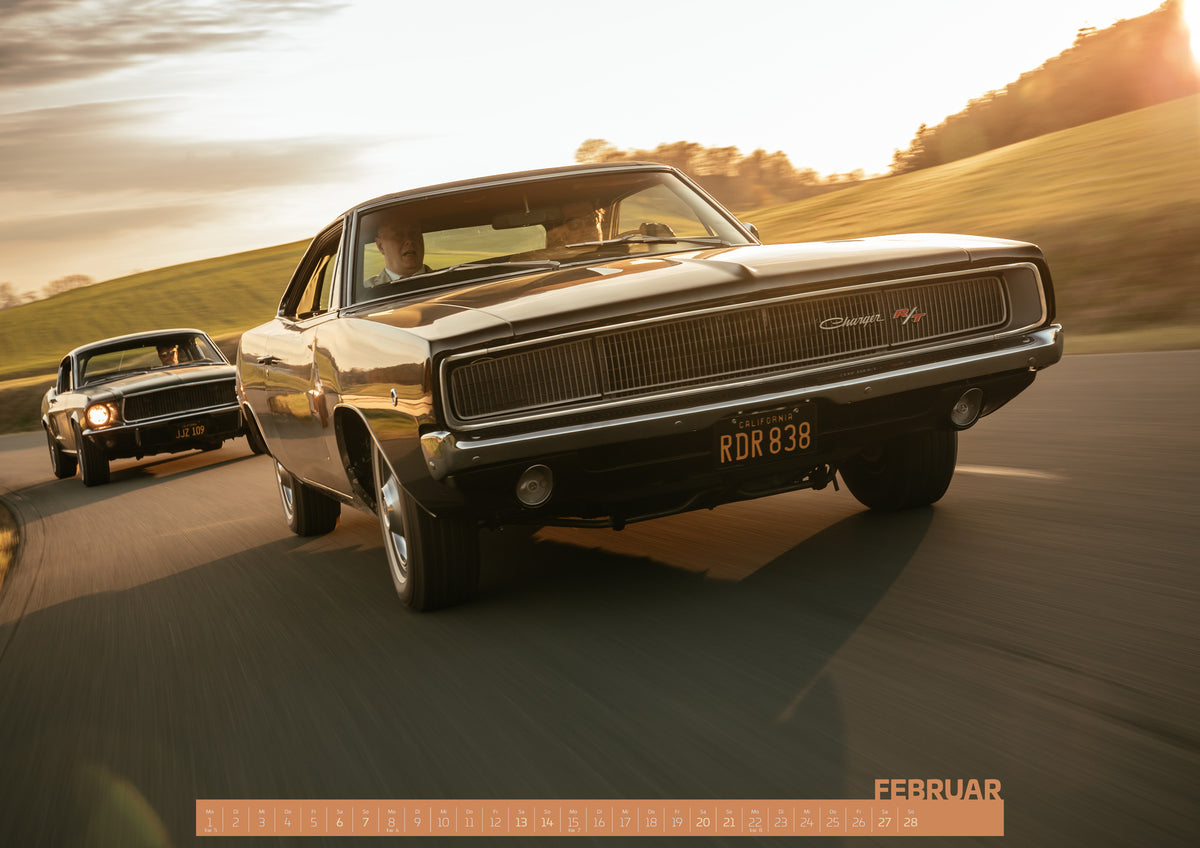 Bullitt Kalender 2027 – Februar: Dunkel erscheinender Dodge Charger R/T aus tiefer Frontperspektive auf einer kurvigen Landstraße; dahinter folgt ein weiteres dunkles Coupé. Goldenes Gegenlicht, grüne Hügel und…