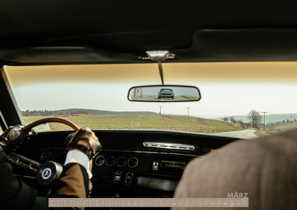 Bullitt Kalender 2027 – März: Blick aus einem dunklen Fahrzeuginnenraum: Behandschuhte Hände am Lenkrad, eine Person rechts und ein dunkler Dodge Charger frontal im Rückspiegel. Vorne verläuft eine helle…
