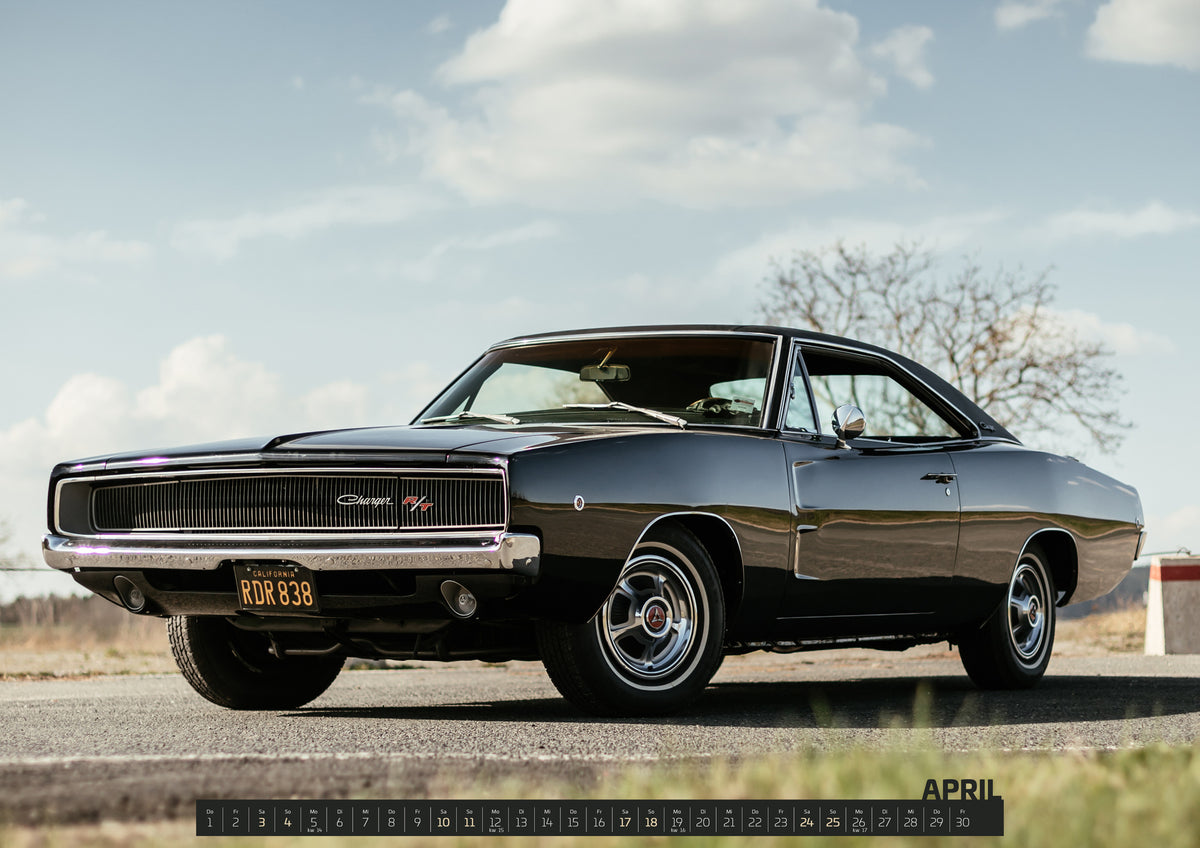 Bullitt Kalender 2027 – April: Dunkler Dodge Charger R/T in tiefer Dreiviertelansicht von vorn auf einer Straße unter hellem, wolkigem Himmel; im Hintergrund kahle Bäume und trockenes Gras