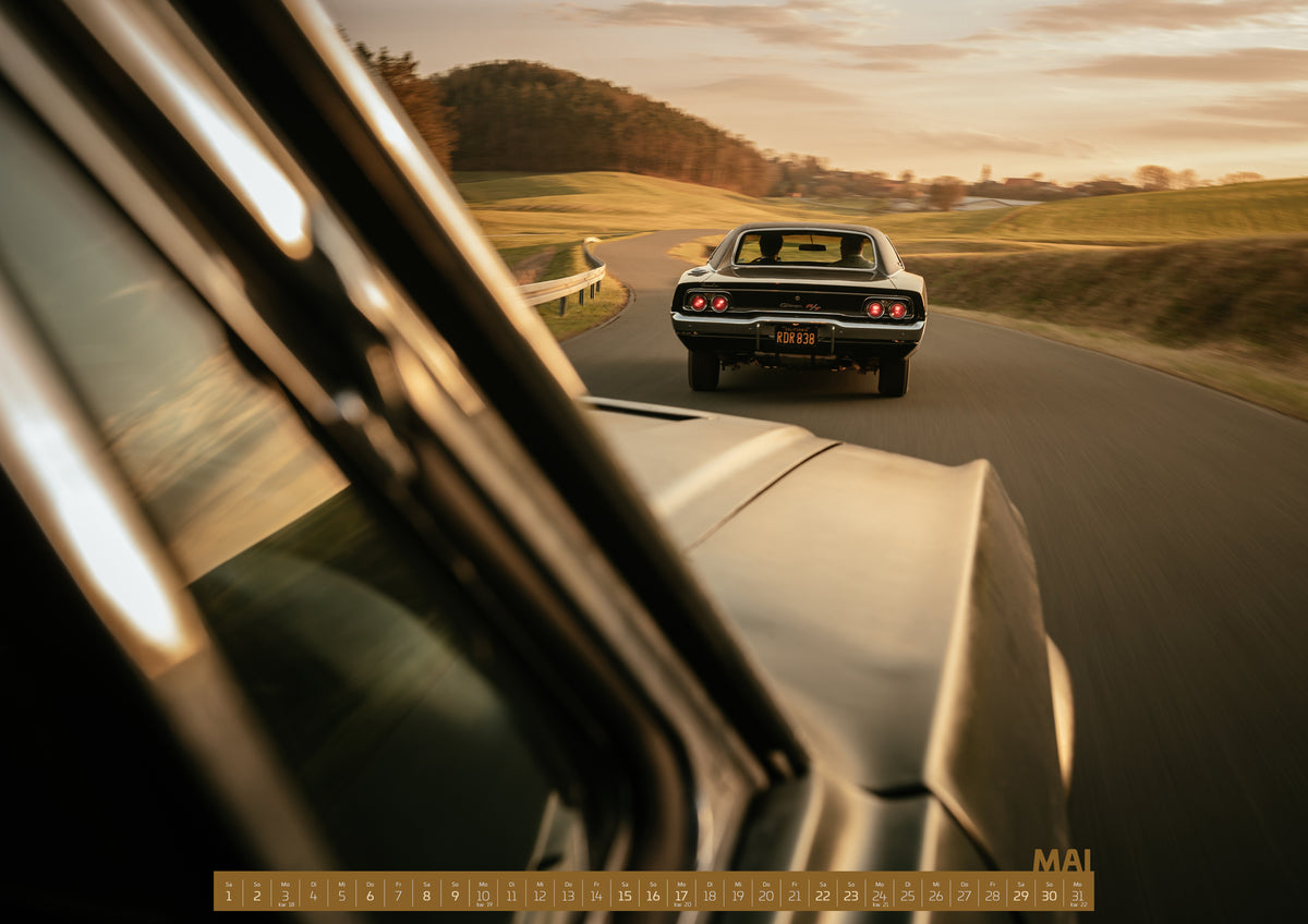 Bullitt Kalender 2027 – Mai: Dunkler Dodge Charger 440 R/T von hinten auf einer kurvigen Landstraße im goldenen Abendlicht, fotografiert aus einem zweiten fahrenden Auto; rote Rückleuchten, Wiesen und…