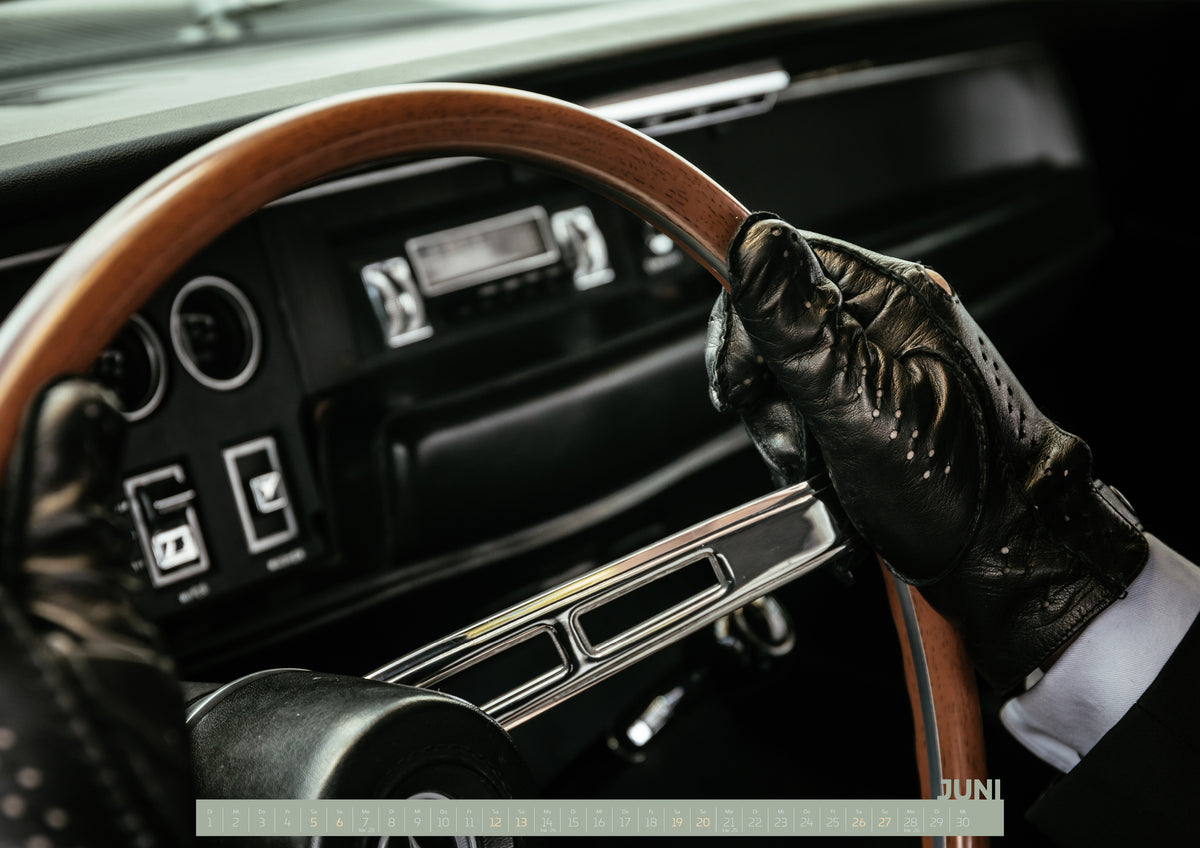 Bullitt Kalender 2027 – Juni: Nahaufnahme eines dunklen Fahrzeugcockpits: Eine Hand in schwarzem, perforiertem Handschuh greift ein braunes, holzfarbenes Lenkrad. Rundinstrumente, Radio und Armaturenbrett liegen…