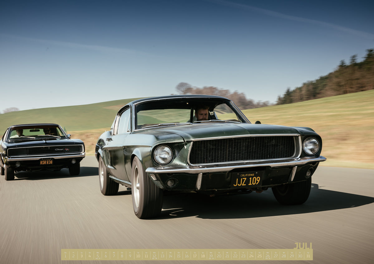 Bullitt Kalender 2027 – Juli: Dynamische Fahraufnahme zweier klassischer Coupés auf einer kurvigen Landstraße. Im Vordergrund fährt ein dunkelgrünes Coupé in Front-Dreiviertelansicht; links dahinter folgt der…