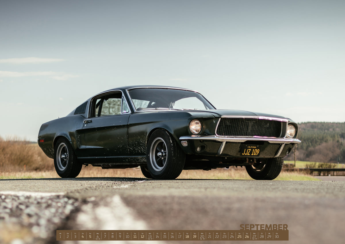 Bullitt Kalender 2027 – September: Dunkelgrüner Ford Mustang in niedriger Dreiviertelansicht von vorn auf Asphalt; dahinter offene Felder, ein bewaldeter Horizont und heller Himmel bei Tageslicht