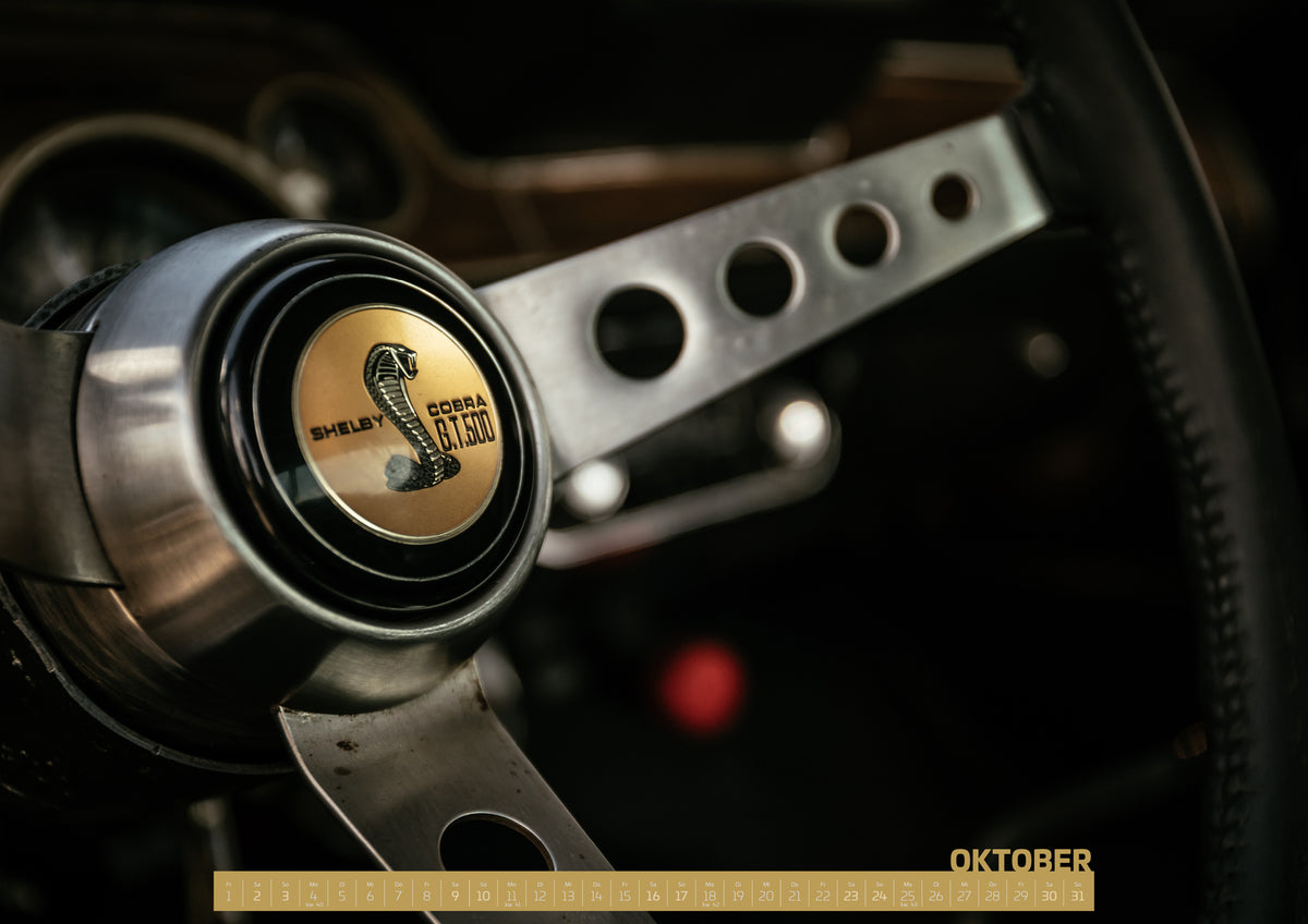 Bullitt Kalender 2027 – Oktober: Nahaufnahme eines dreispeichigen Lenkrads mit gelochten silberfarbenen Speichen, schwarzem Kranz und goldenem Shelby-Cobra-GT500-Emblem in einem dunklen, warm beleuchteten Cockpit