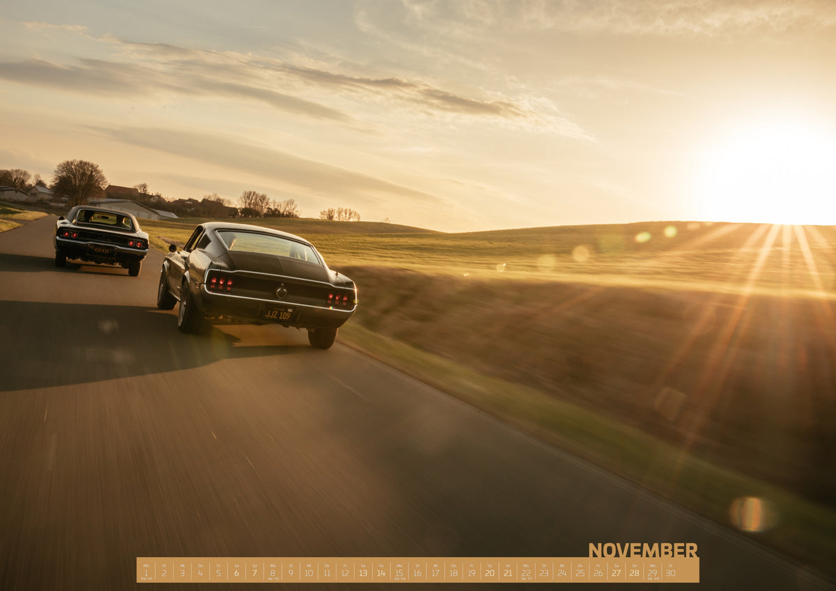 Bullitt Kalender 2027 – November: Zwei dunkel erscheinende klassische Coupés von hinten auf einer kurvigen Landstraße; eines fährt links voraus, das zweite füllt den mittleren Vordergrund. Tief stehende Sonne rechts…