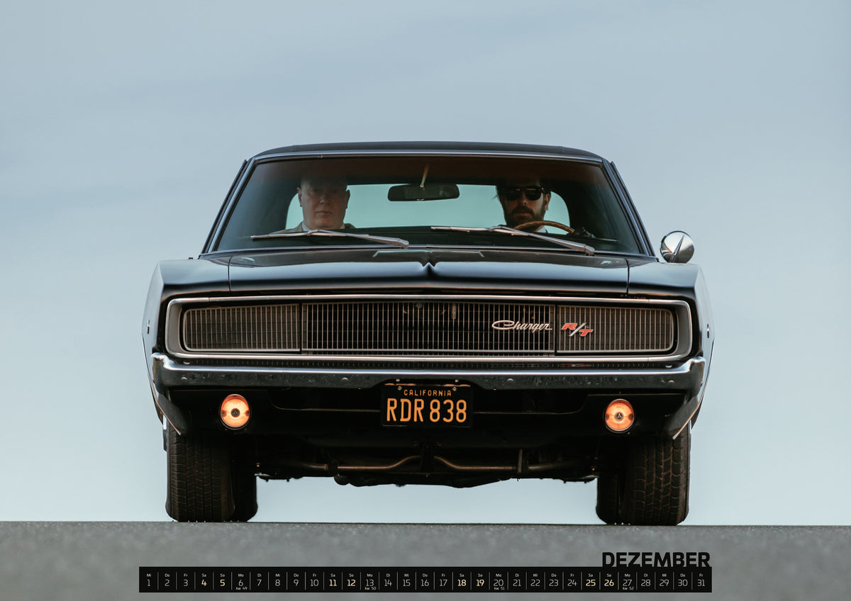 Bullitt Kalender 2027 – Dezember: Sehr dunkler Dodge Charger R/T in tiefer, zentraler Frontalansicht vor blassblauem Himmel; zwei Personen sitzen hinter der Windschutzscheibe, zwei Zusatzleuchten glimmen warm