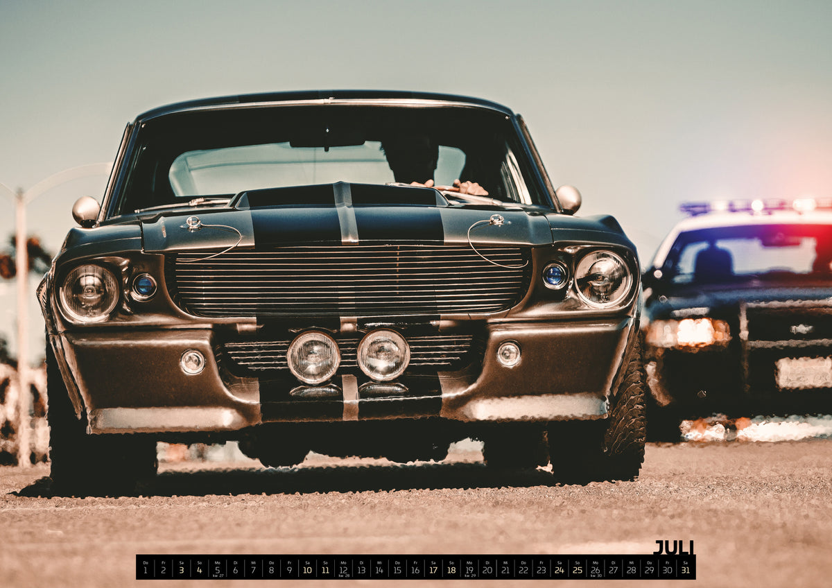 Eleanor Kalender 2027 – Juli: Dunkler Eleanor Ford Mustang Shelby GT500 in tiefer Frontalansicht auf heller Fahrbahn, mit dunklen Längsstreifen, Motorhaubenaufsatz, Rundscheinwerfern und zwei Zusatzleuchten…