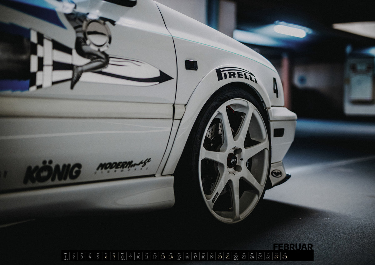 Jesse’s Jetta Kalender 2027 – Februar: Ein Ausschnitt von Jesses Jetta zeigt weißes Seitenteil, graue Grafik, Vorderrad und Motorsport-Schriftzüge in einer Werkstatt