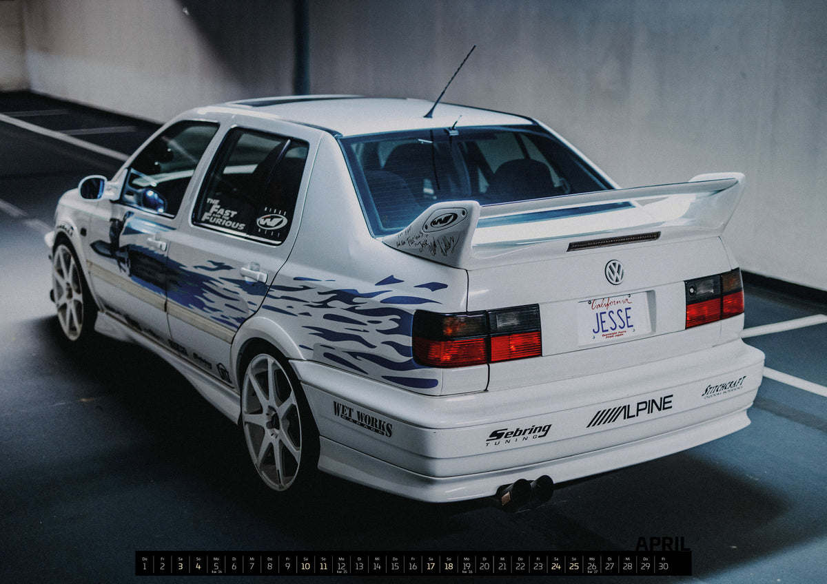 Jesse’s Jetta Kalender 2027 – April: Weißer VW Jetta in rückwärtiger Dreiviertelansicht in einem Parkbereich, mit blauen Flammengrafiken, großem Heckflügel, hellen Mehrspeichenrädern und roten Rückleuchten bei kühlem Licht