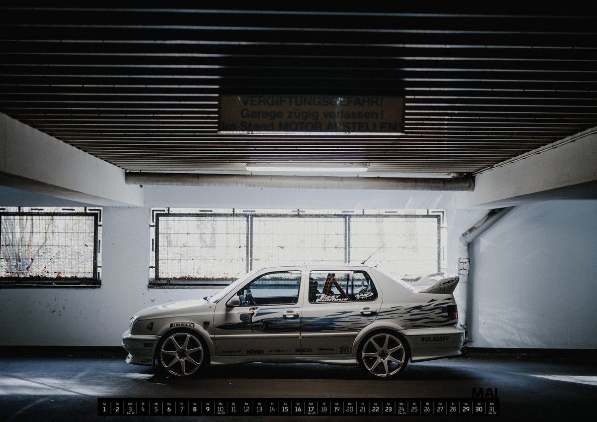 Jesse’s Jetta Kalender 2027 – Mai: Silberfarbener VW Jetta mit blau-schwarzer Seitengrafik, großen Leichtmetallrädern und hohem Heckflügel im Seitenprofil in einer dunklen Parkgarage