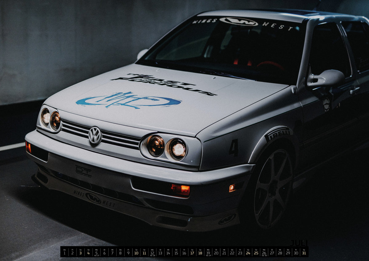 Jesse’s Jetta Kalender 2027 – Juli: Heller Volkswagen Jetta in frontaler Dreiviertelansicht vor dunklem Hintergrund, mit eingeschalteten runden Frontscheinwerfern, orangefarbenen Leuchten, schwarzen und blauen Grafiken…