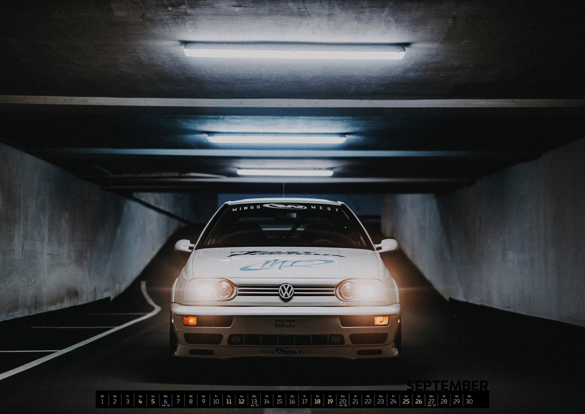 Jesse’s Jetta Kalender 2027 – September: Weißer Volkswagen Jetta in frontaler Ansicht in einer dunklen Betongarage; Scheinwerfer und orangefarbene untere Leuchten sind eingeschaltet, darüber verlaufen helle Deckenröhren