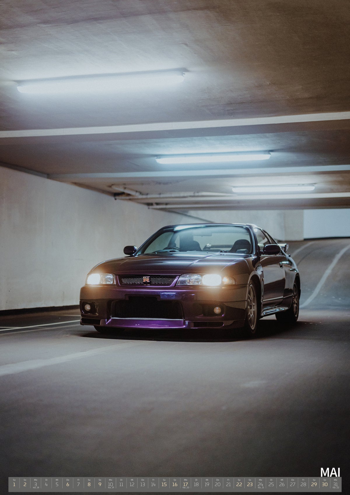 Midnight Purple I Kalender 2027 – Mai: Der Nissan Skyline R33 steht klein und frontal am Ende einer dunklen Parkhausspur; ein warmer Scheinwerfer bricht das kühle Umfeld