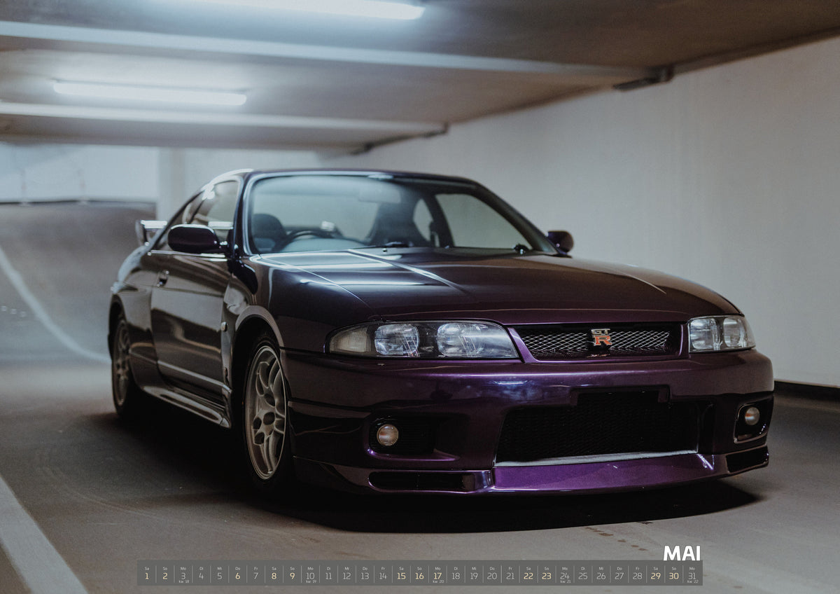 Midnight Purple I Kalender 2027 – Mai: Der Nissan Skyline R33 steht tief in einer Dreiviertel-Frontansicht zwischen weißen Fahrbahnmarkierungen und kühlen Betonflächen