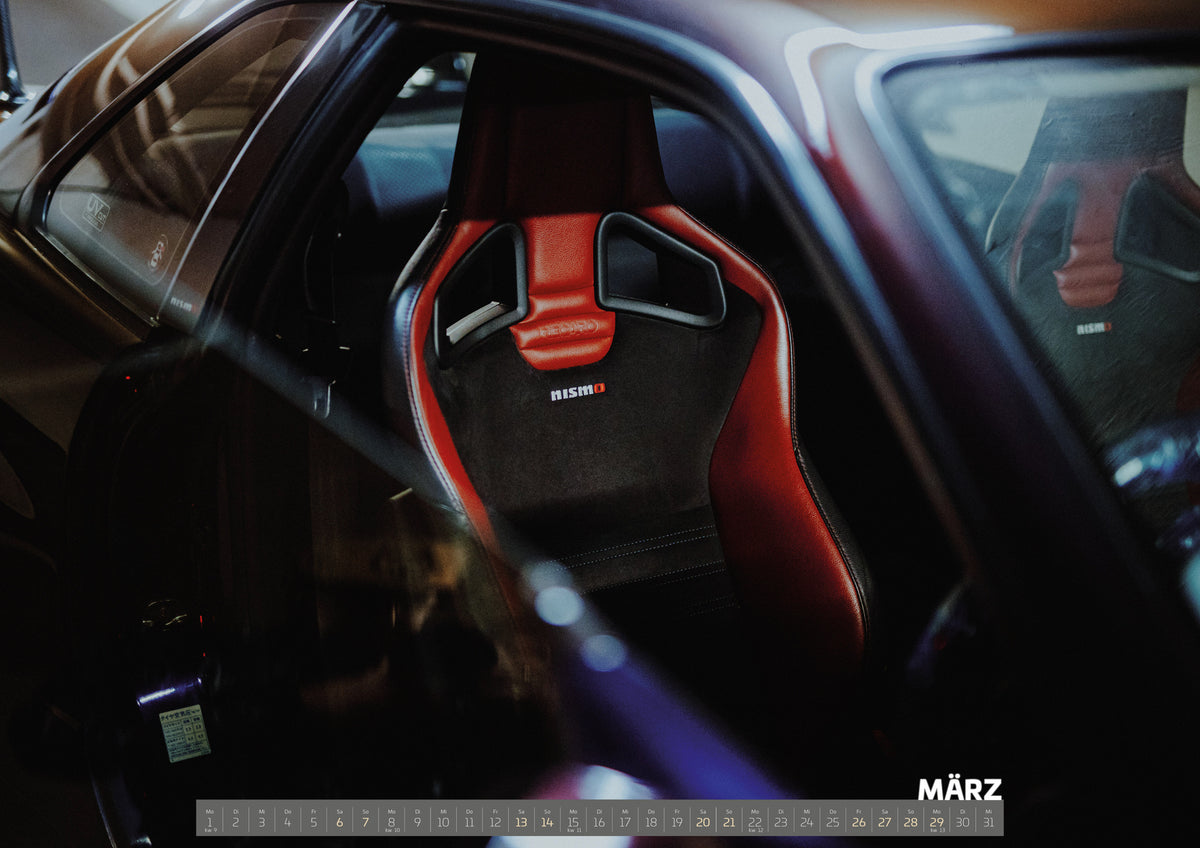 Midnight Purple Collection Kalender 2027 – März: Seitlicher Nahblick in den dunklen Innenraum eines Nissan Skyline R34. Ein rot-schwarzer Sportsitz mit Öffnungen in der Rückenlehne und Nismo-Schriftzug steht im Zentrum; ein zweiter…
