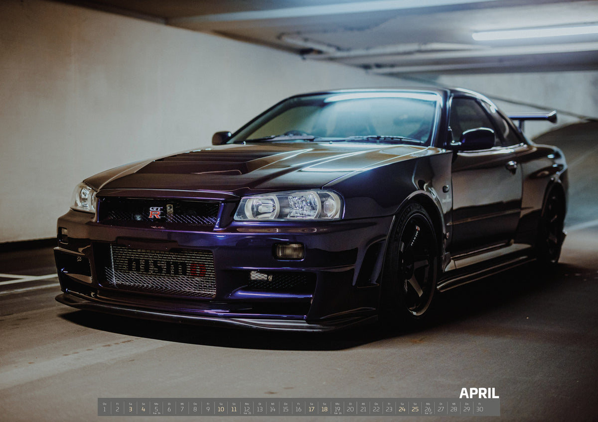Midnight Purple Collection Kalender 2027 – April: Dunkelvioletter Nissan Skyline R34 in Front-Dreiviertelansicht auf einem Parkdeck, mit schwarzem Kühlergrill, dunklen Rädern und großem Heckflügel. Helle Deckenleuchten spiegeln sich…