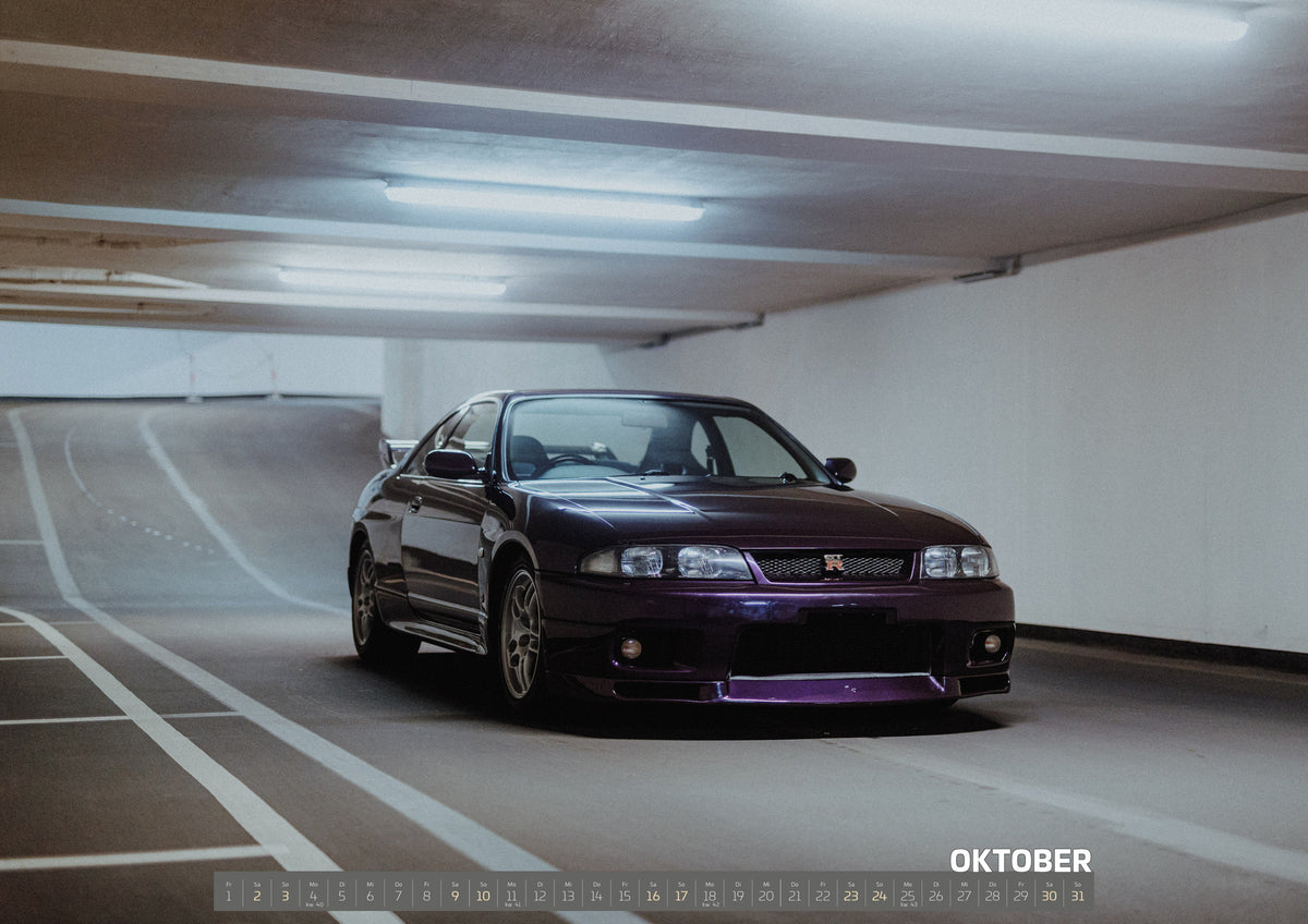 Midnight Purple Collection Kalender 2027 – Oktober: Der Nissan Skyline R33 steht in einer breiten Dreiviertelansicht zwischen weißen Fahrbahnlinien; violette Lackflächen glühen im Neonlicht
