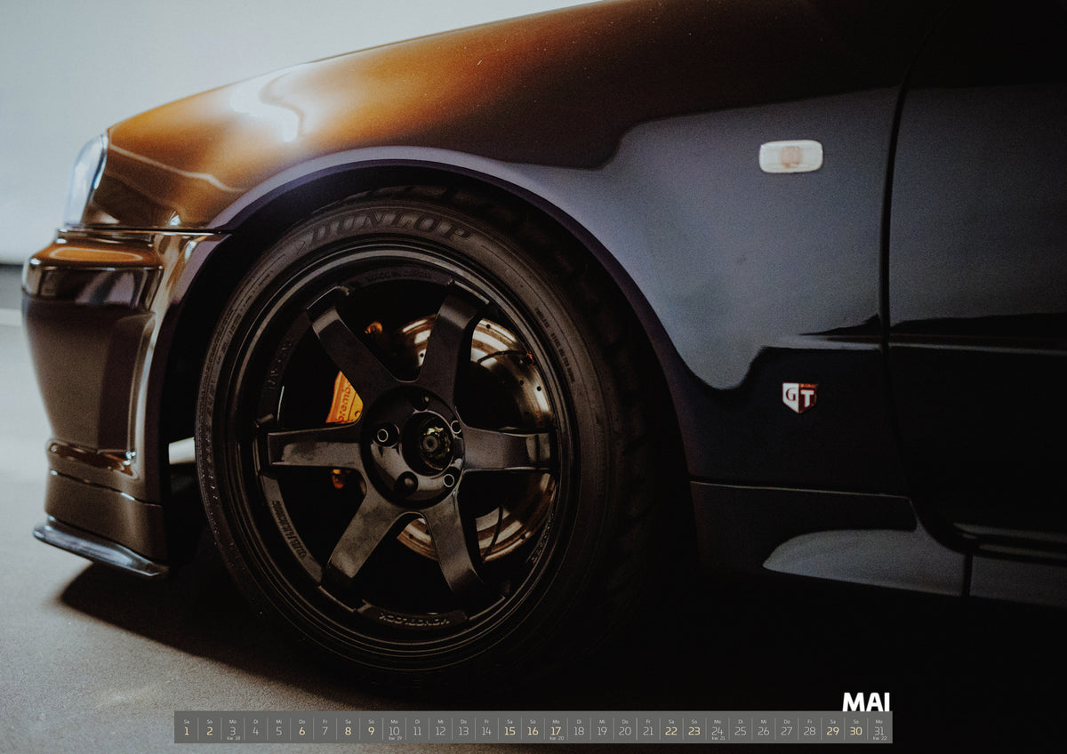 R34 Midnight Purple II Kalender 2027 – Mai: Die Nahaufnahme konzentriert sich auf Vorderrad, Bremse und Kotflügel des Nissan Skyline R34; der violette Lack spiegelt das Parkhauslicht