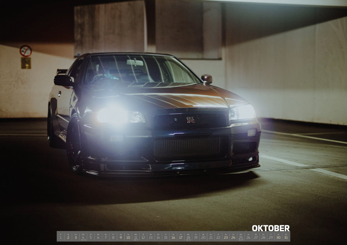R34 Midnight Purple II Kalender 2027 – Oktober: Der Nissan Skyline R34 kommt frontal aus dem dunklen Parkhaus; ein kräftiger Scheinwerferreflex dominiert die Szene