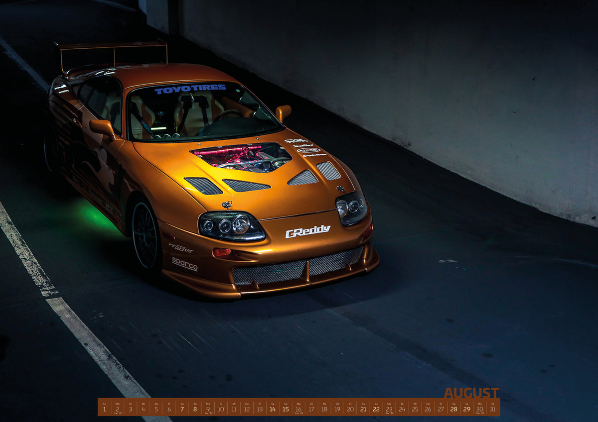 ChromeCars Retrospektiv II Kalender 2027 – August: Slap Jacks orange Toyota Supra ist aus erhöhter Perspektive in einer dunklen Halle aufgenommen; grüne Unterbodenbeleuchtung setzt einen Neonakzent