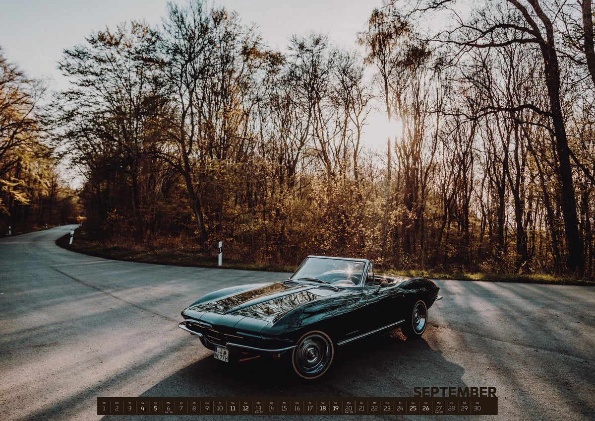 ChromeCars Retrospektiv II Kalender 2027 – September: Eine dunkelgrüne 1967 Chevrolet Corvette C2 als offenes Cabriolet in Front-Dreiviertelansicht auf einer kurvigen Waldstraße. Tief stehende Sonne scheint durch kahle Bäume; Gegenlicht…