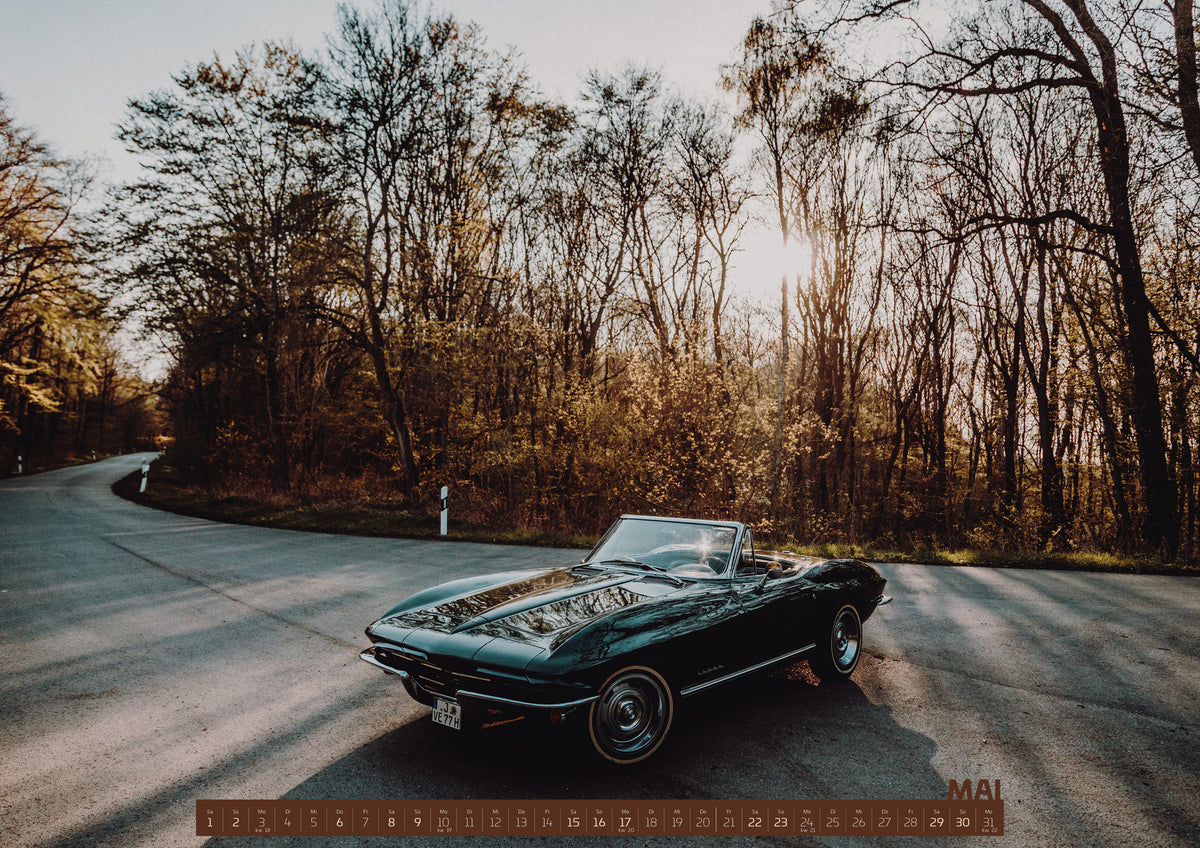ChromeCars Retrospektiv Kalender 2027 – Mai: Eine dunkelgrüne 1967 Chevrolet Corvette C2 als offenes Cabriolet in Front-Dreiviertelansicht auf einer kurvigen Waldstraße. Tief stehende Sonne scheint durch kahle Bäume; Gegenlicht…
