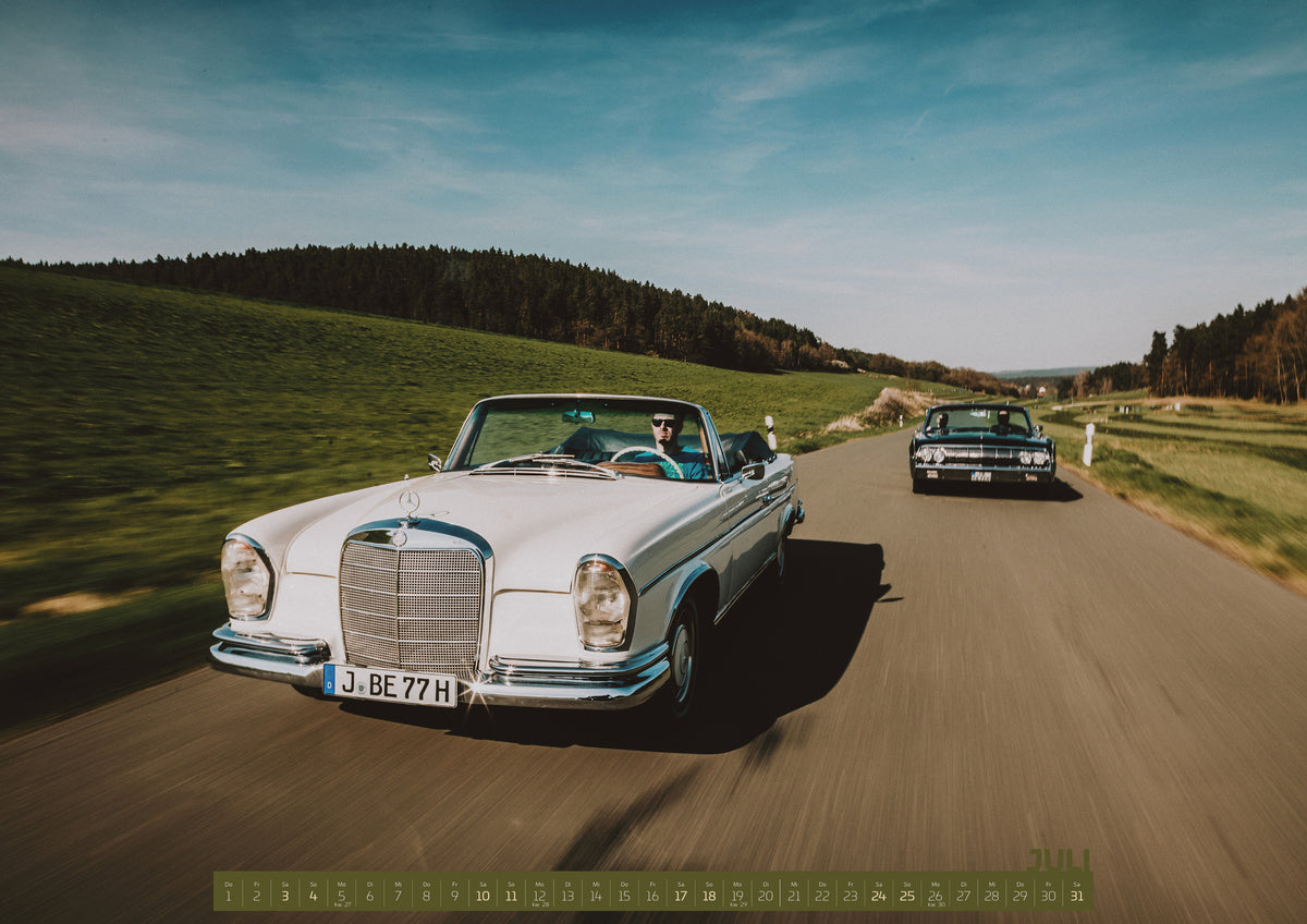 ChromeCars Retrospektiv Kalender 2027 – Juli: Frontale Fahraufnahme eines weißen 1965 Mercedes 300 SE W112 Cabriolets im Vordergrund und eines dunklen 1964 Lincoln Continental Cabriolets dahinter auf einer Landstraße. Grüne…