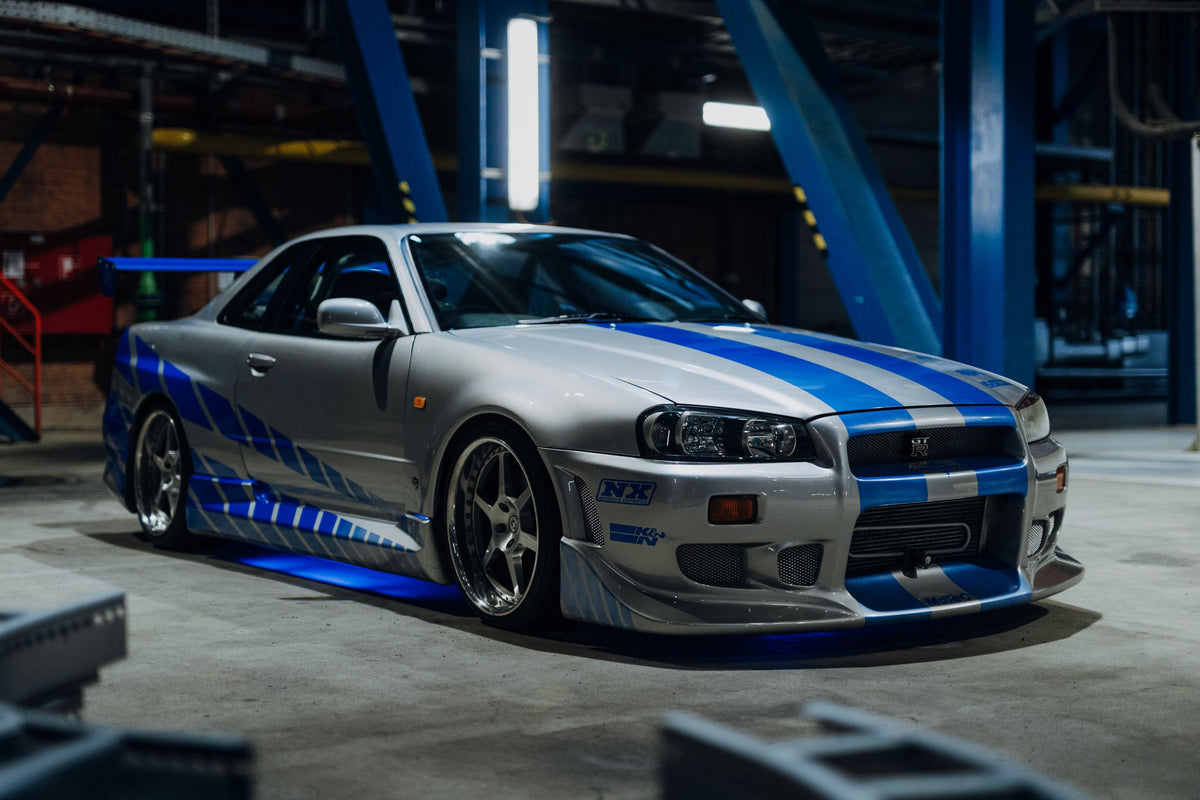 Silberner Nissan Skyline GT-R R34 mit blauen Racing-Streifen und blauer Unterbodenbeleuchtung in einer Industriehalle.