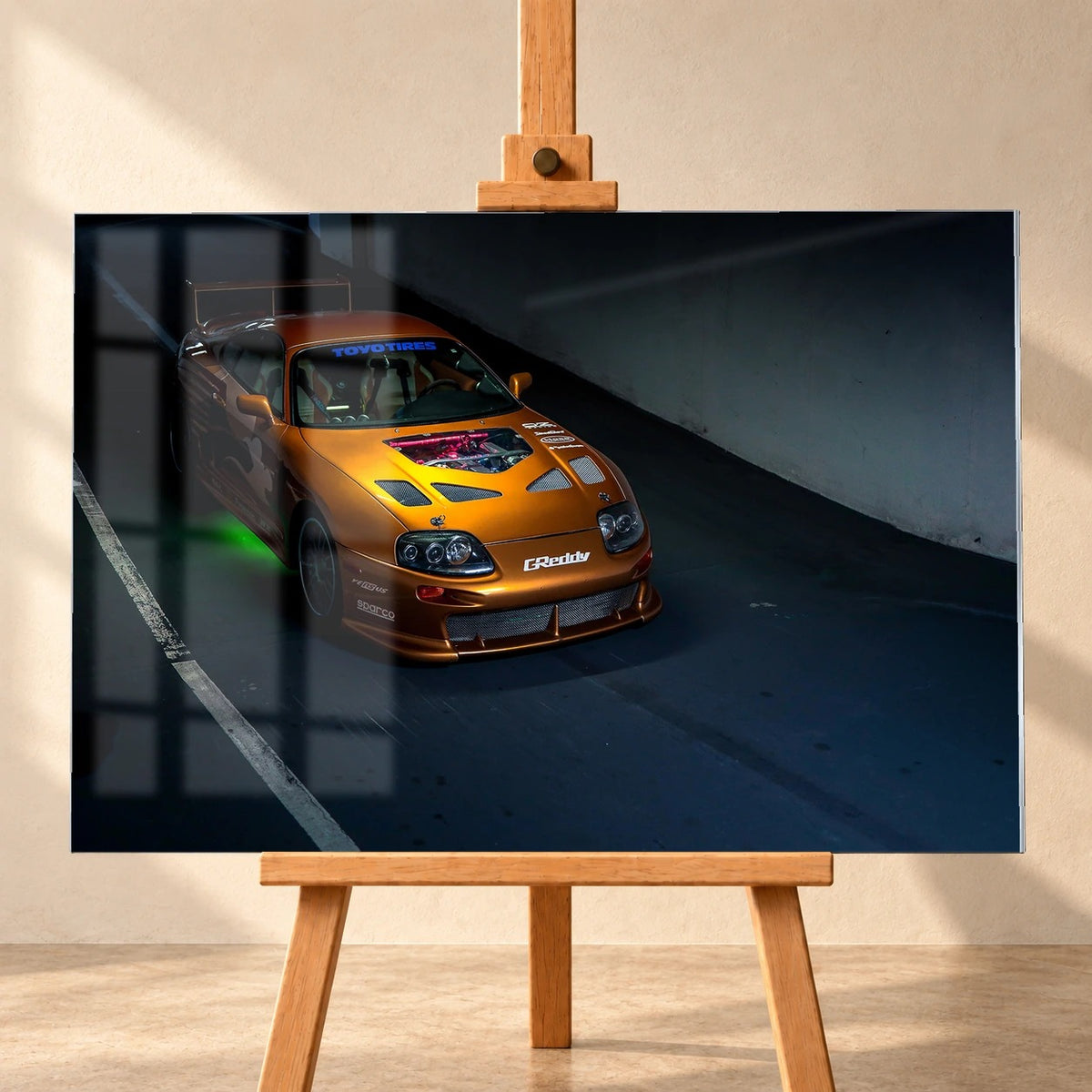 Frontale Produktvisualisierung eines Acrylglasbilds auf einer Holzstaffelei vor heller Wand. Eine Fensterspiegelung überlagert links das Motiv des orangefarbenen Toyota Supra aus „2 Fast 2 Furious“, der schräg von oben und vorn mit grüner Unterbodenbeleuchtung zu sehen ist.