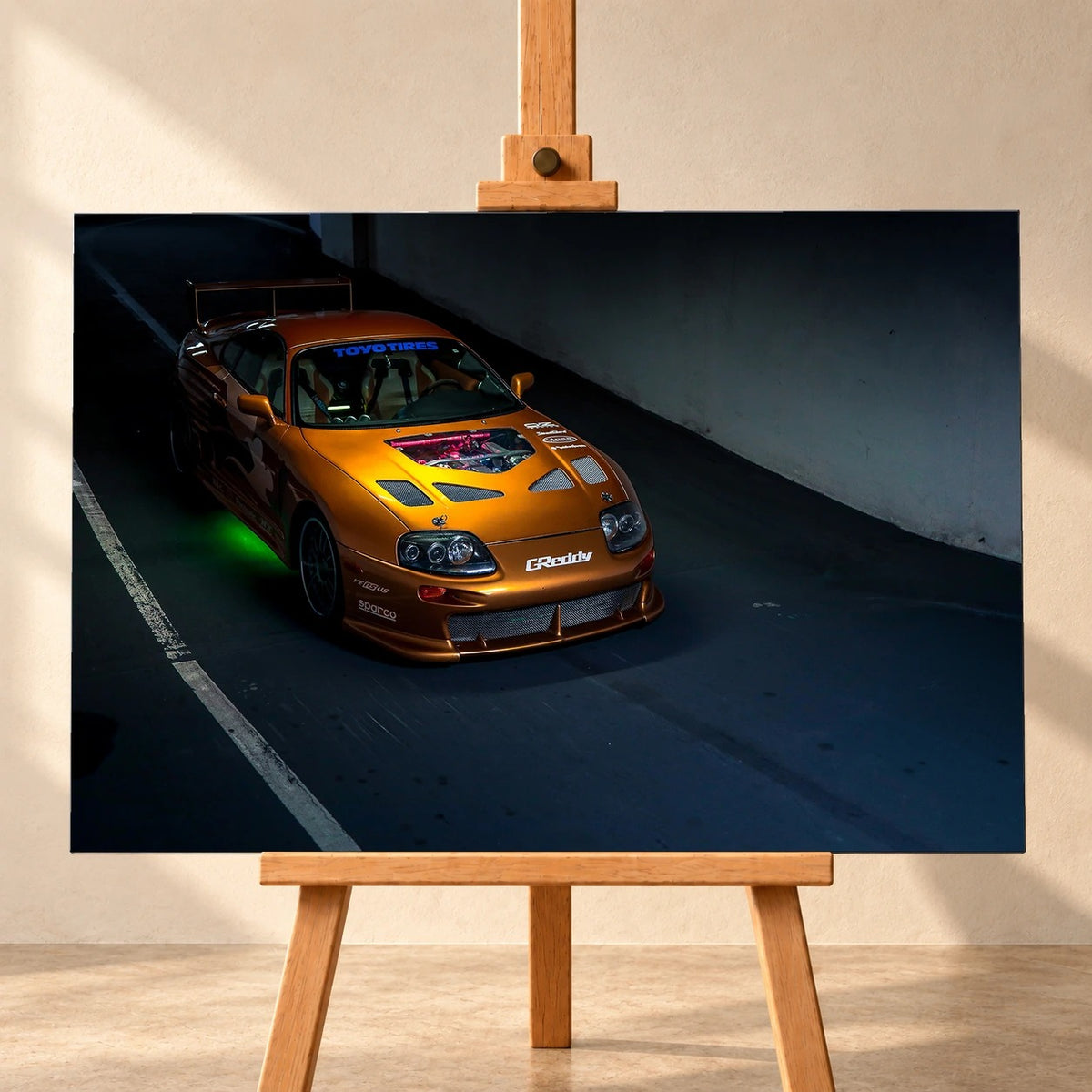 Visualisierung eines querformatigen Leinwandbilds auf einer Holzstaffelei vor heller Wand. Das Motiv zeigt den orangefarbenen Toyota Supra aus „2 Fast 2 Furious“ schräg von oben und vorn, mit großem Heckflügel und grüner Unterbodenbeleuchtung.