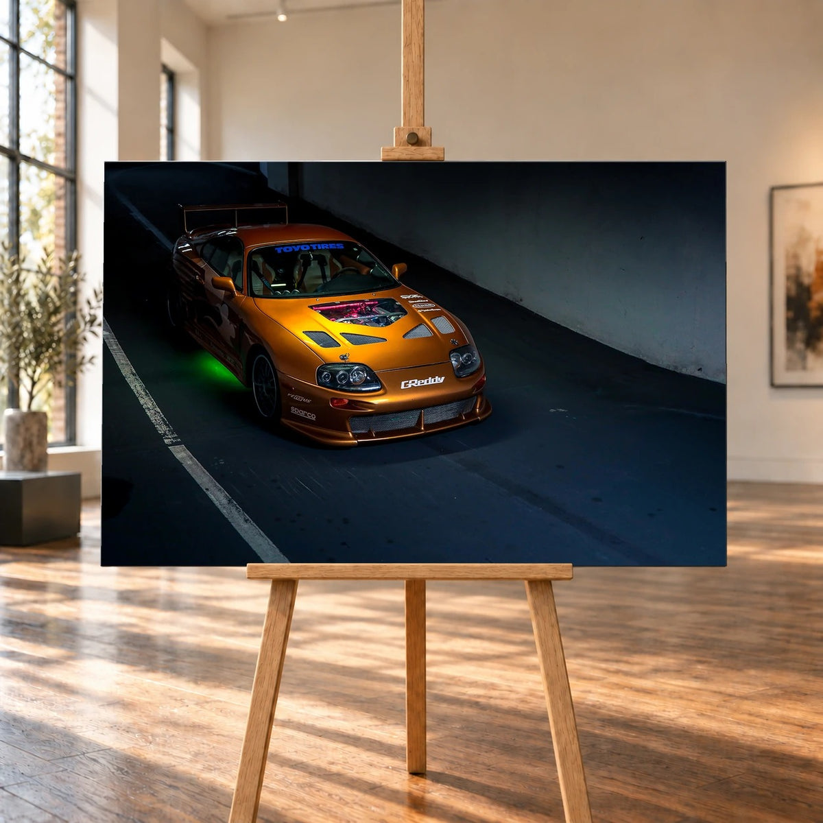 Raumvisualisierung mit einem querformatigen Leinwandbild auf einer Holzstaffelei zwischen hohen Fenstern und sonnenbeschienenem Holzboden. Das frontal gezeigte Bild trägt das Motiv des orangefarbenen Toyota Supra aus „2 Fast 2 Furious“ mit Heckflügel und grüner Unterbodenbeleuchtung in dunkler Umgebung.