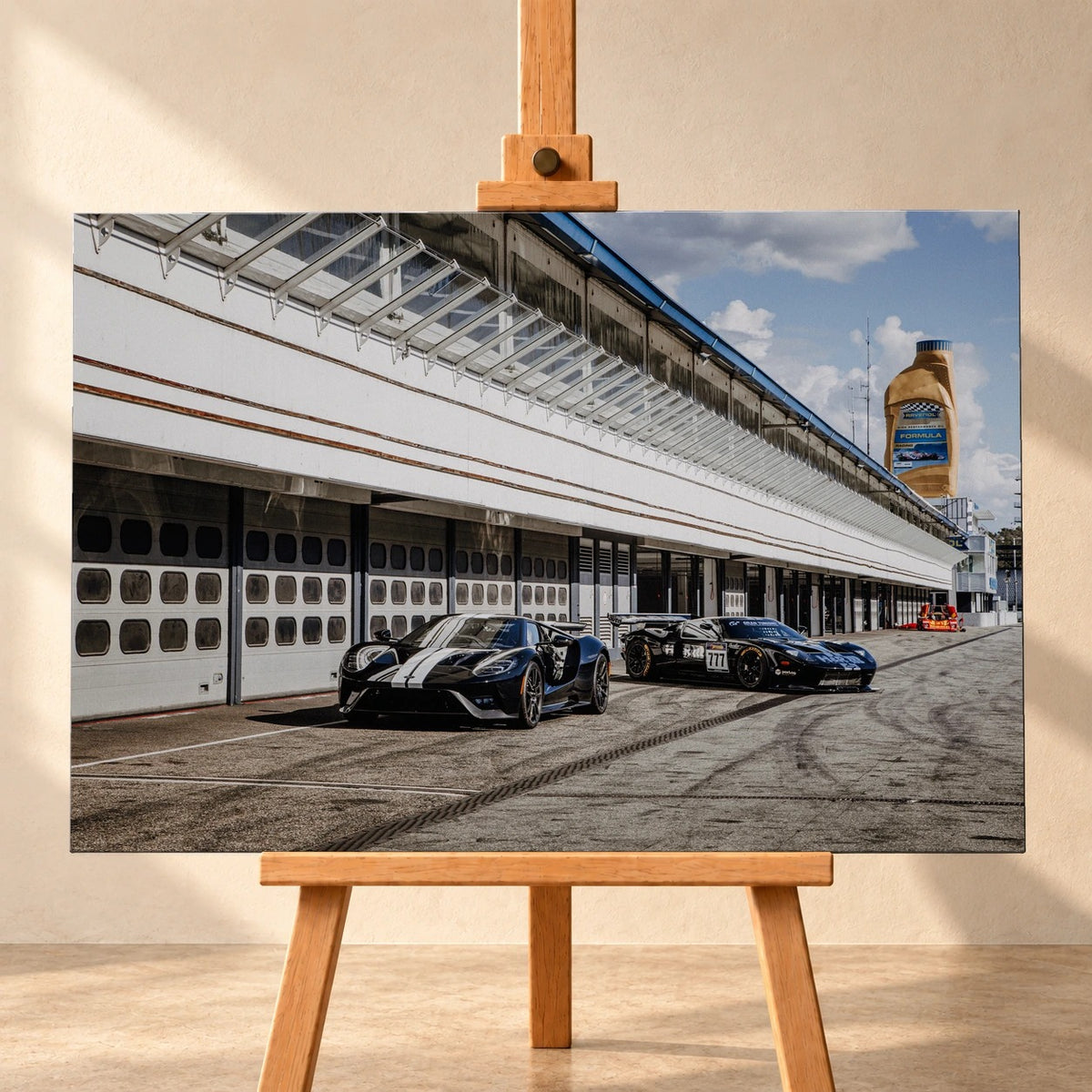 Frontale Visualisierung eines querformatigen Leinwandbildes auf einer Holzstaffelei vor heller Wand. Das Motiv zeigt einen schwarzen Ford GT mit weißen Längsstreifen neben einem Ford GT GT3 mit Startnummer 777 in einer Boxengasse.
