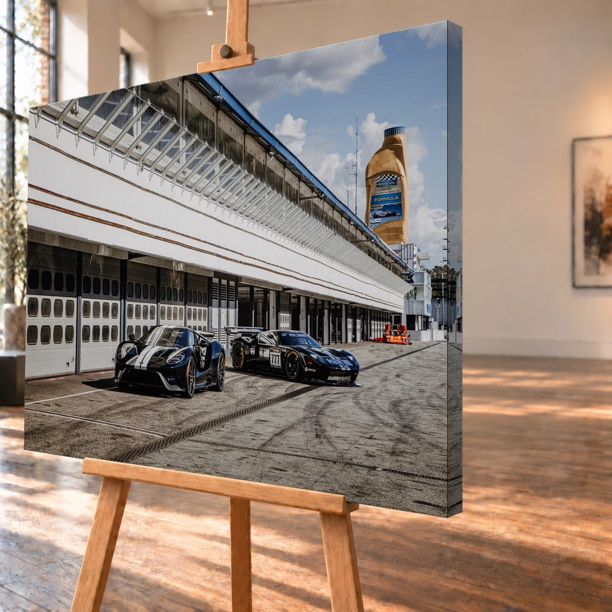 Schrägansicht eines als Leinwandbild visualisierten Motivs mit Ford GT und Ford GT GT3 vor einer langen Boxenanlage. Das Bild steht auf einer Holzstaffelei; an der sichtbaren rechten Seitenkante setzt sich das Motiv fort.