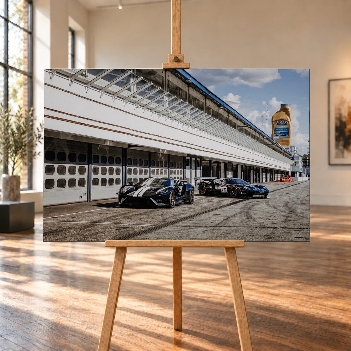 Raumvisualisierung mit einem querformatigen Leinwandbild auf einer Holzstaffelei in einem hellen Raum mit hohen Fenstern und Holzboden. Auf dem Bild stehen ein Ford GT mit weißen Streifen und ein Ford GT GT3 mit Startnummer 777 vor Boxentoren.