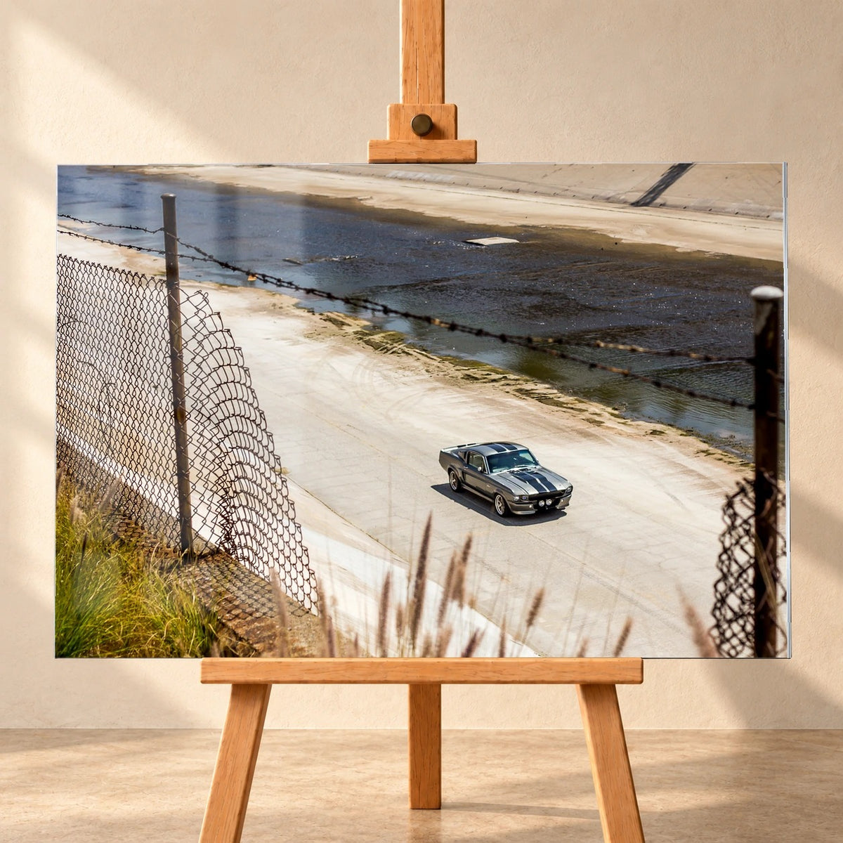 Visualisierung eines querformatigen Acrylglasbilds, frontal auf einer Holzstaffelei vor heller Wand präsentiert. Der silbergraue Ford Mustang Shelby GT500 „Eleanor“ mit dunklen Doppelstreifen ist im Betonkanal zu sehen, ein Drahtzaun liegt im Vordergrund des Motivs.