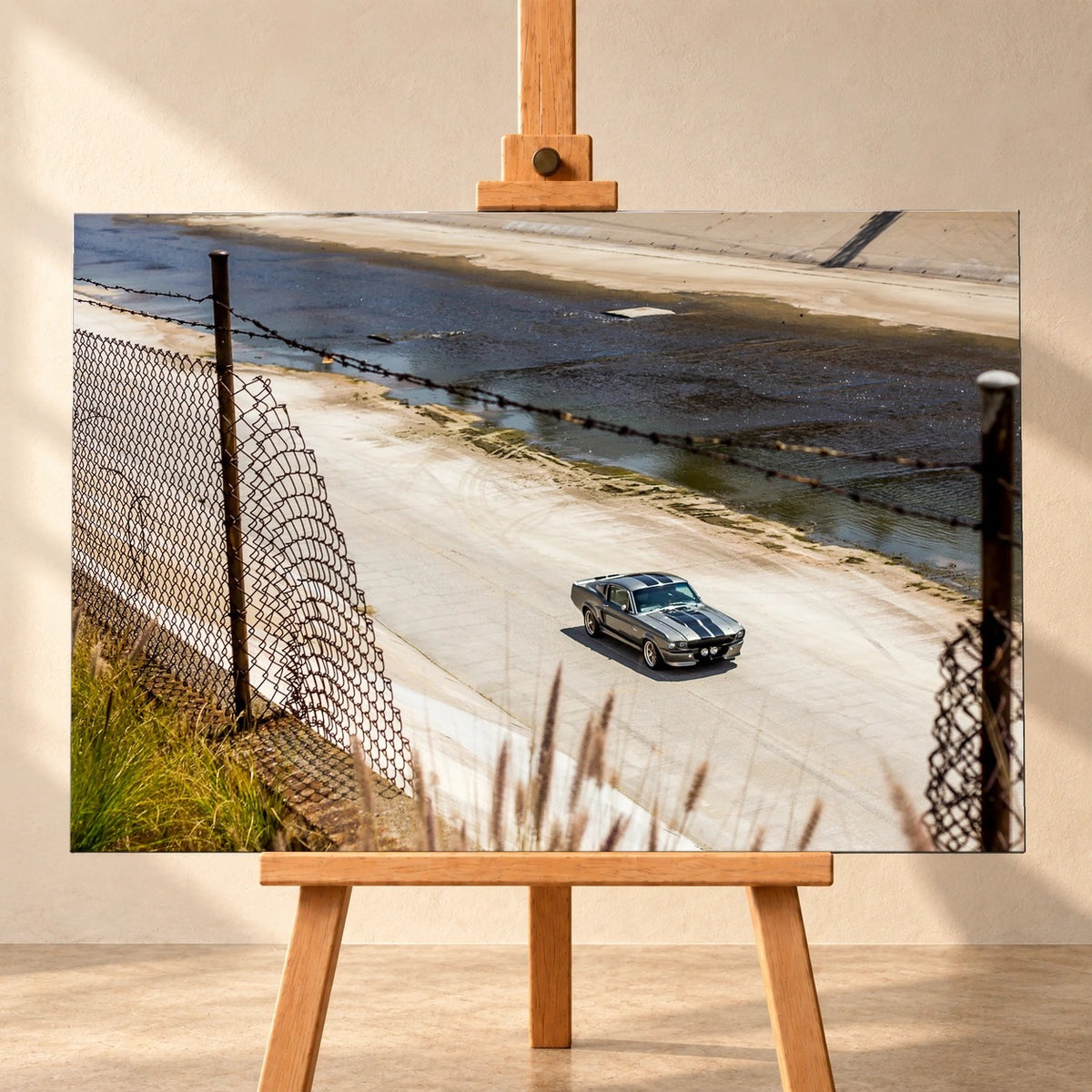 Frontale Visualisierung eines Leinwandbilds im Querformat auf einer Holzstaffelei vor heller Wand. Das Motiv zeigt den silbergrauen Ford Mustang Shelby GT500 „Eleanor“ mit dunklen Streifen in einem Betonkanal hinter einem Drahtzaun.