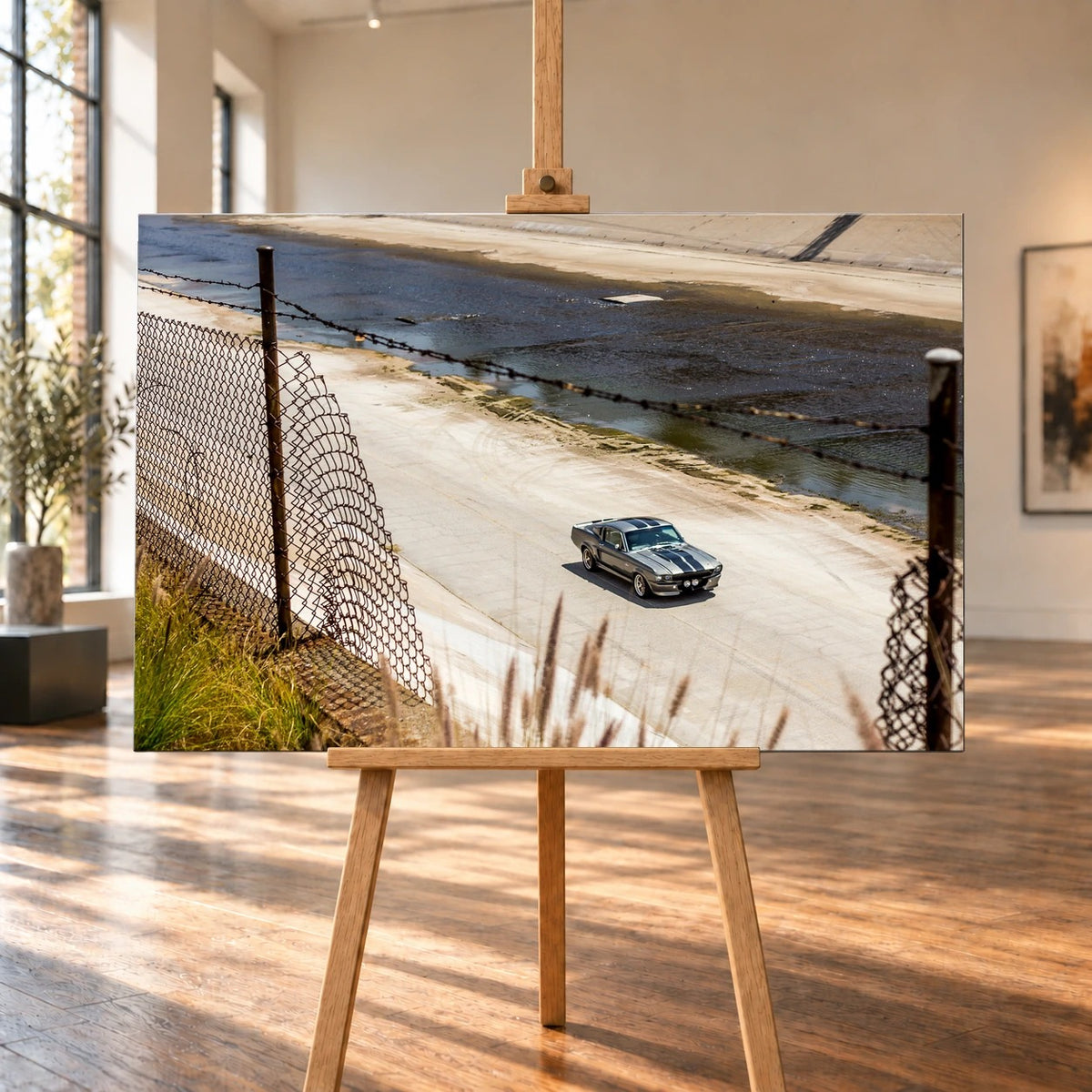 Raumvisualisierung mit einem querformatigen Leinwandbild auf einer Holzstaffelei in einem hellen Raum mit hohen Fenstern und Holzboden. Auf dem Bild ist der silbergraue Ford Mustang Shelby GT500 „Eleanor“ hinter einem Drahtzaun im Betonkanal zu sehen.